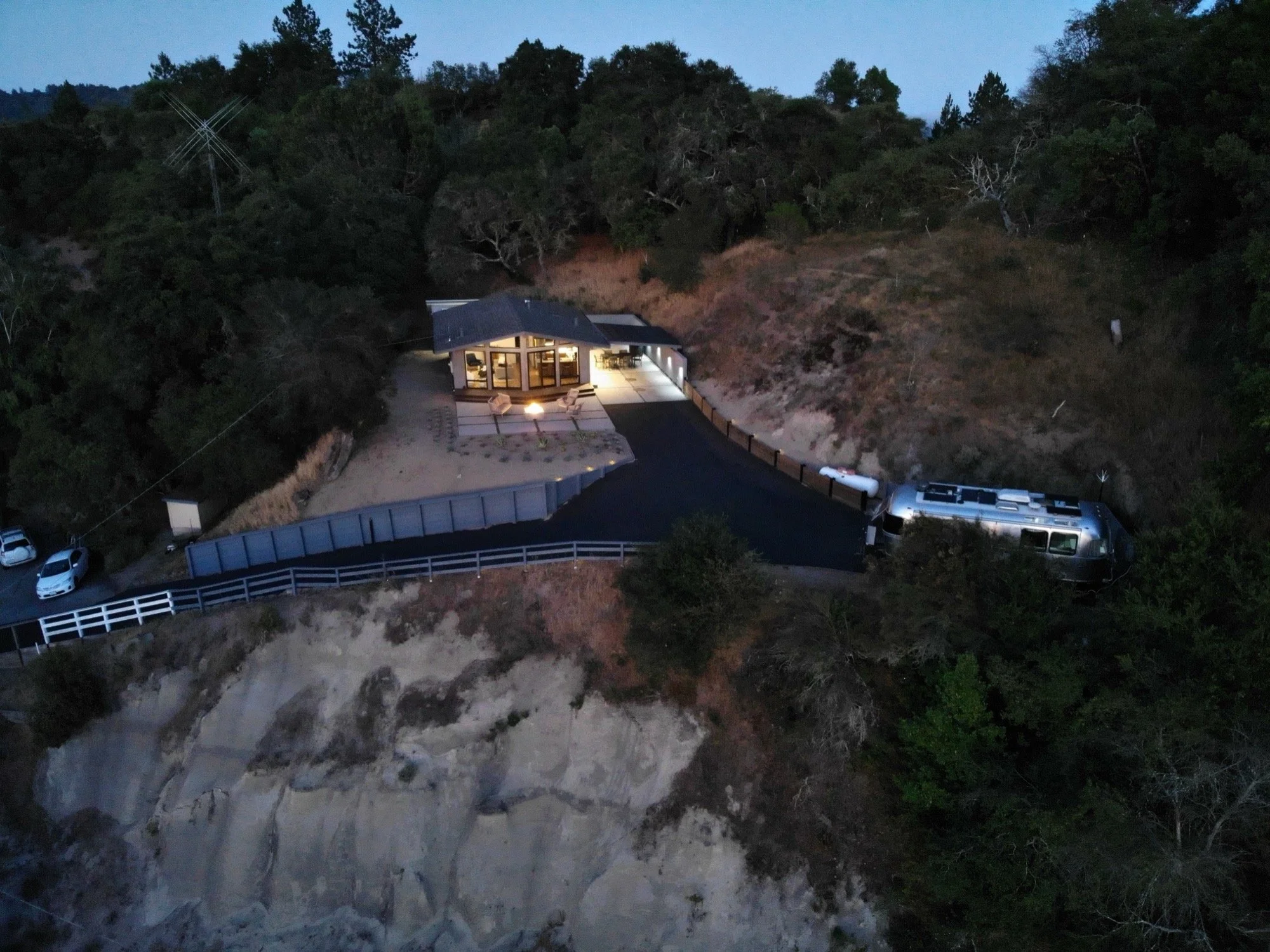 An aerial view of a house built on a hillside with a dark driveway leading to the house, surrounded by trees and rocks, during twilight or early evening.