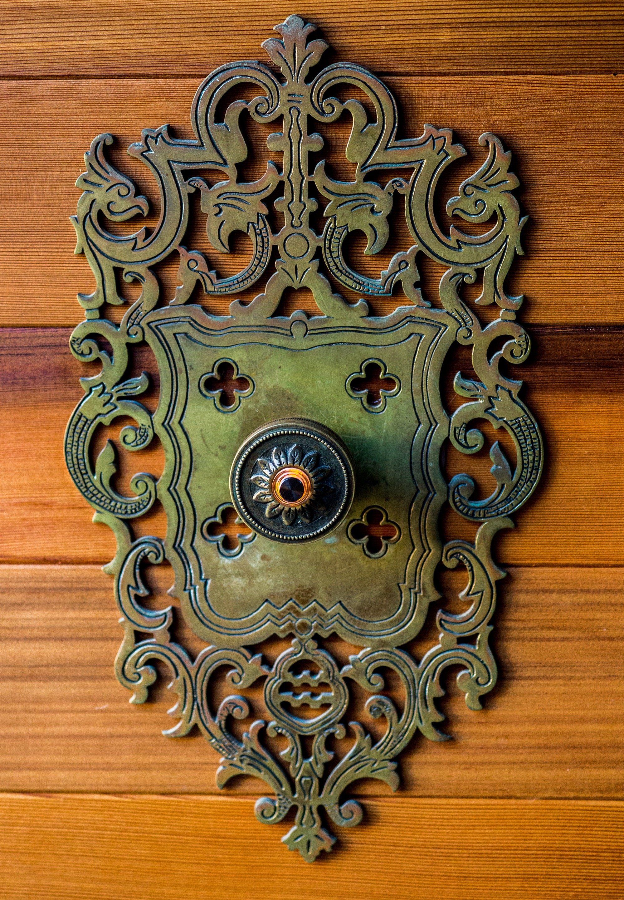 Vintage brass doorbell with ornate decorative design mounted on a wooden wall.