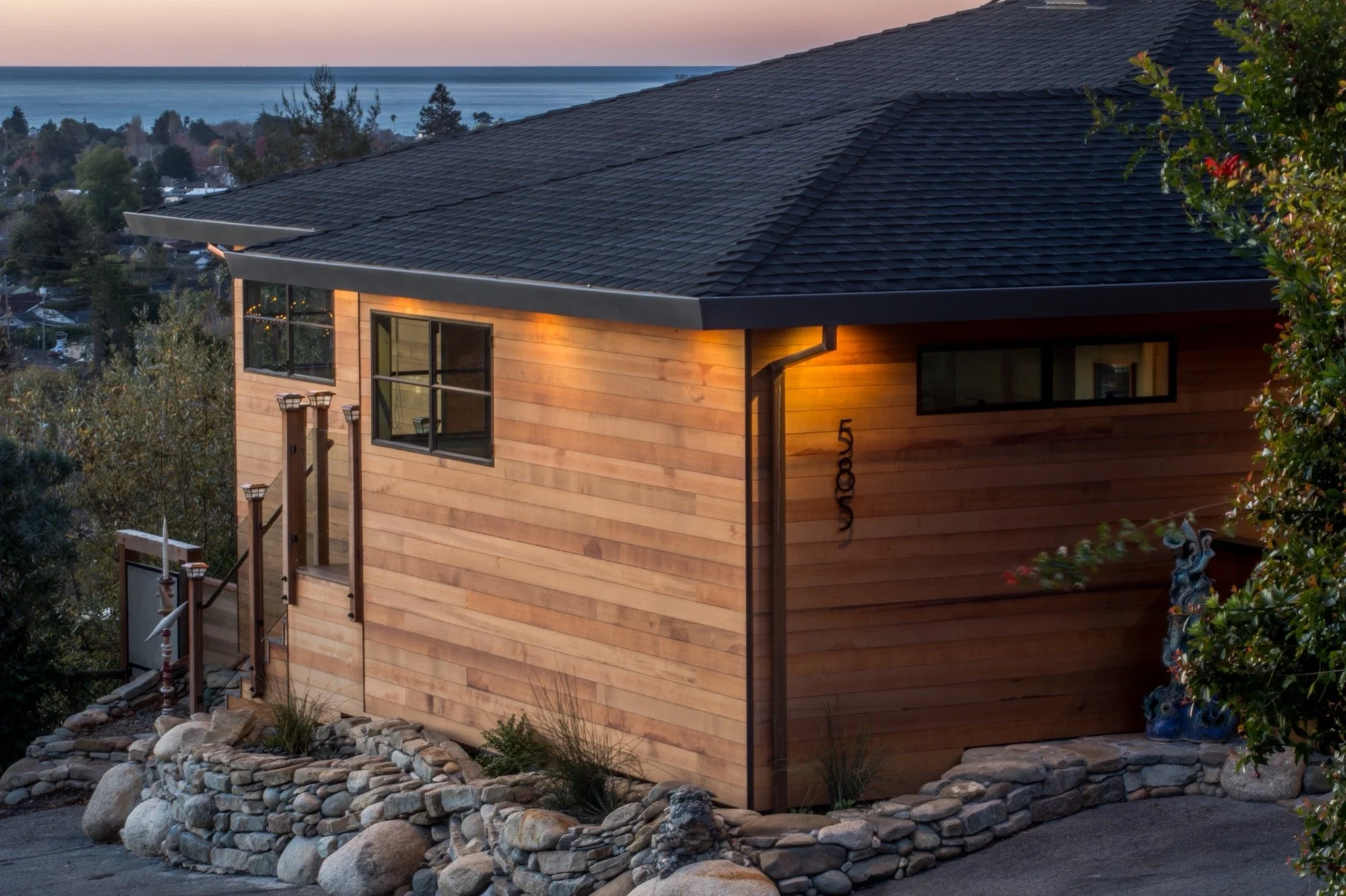 A modern house with a wooden exterior, large windows, and a black shingled roof, situated on a hillside with a stone retaining wall and trees, overlooking a body of water at sunset.