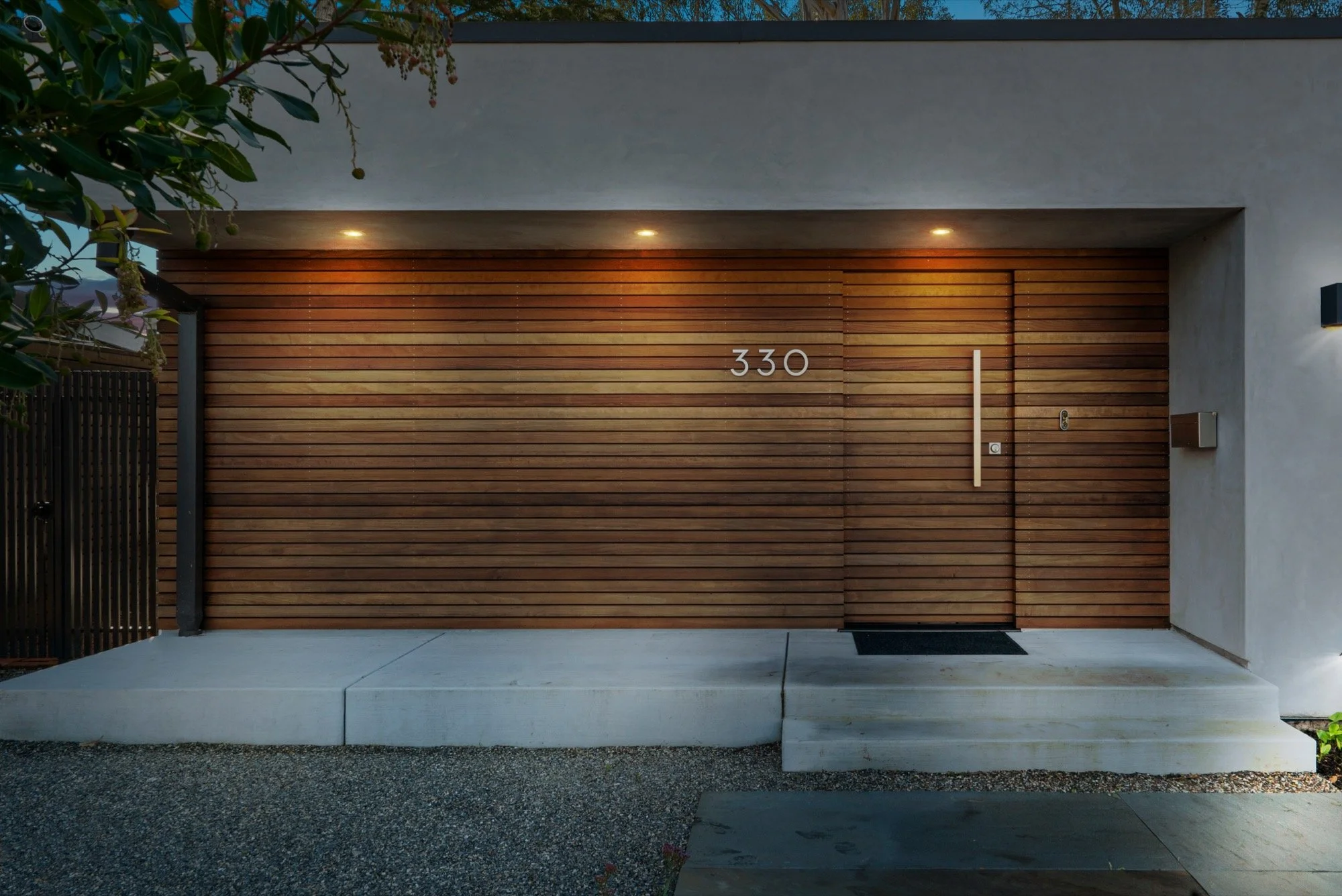 Modern house entrance with a wooden sliding door, house number 330 in silver, and a small doorbell on the wall to the right. The entrance is illuminated by three recessed lights above, and there is a concrete step leading up to the door, with gravel 