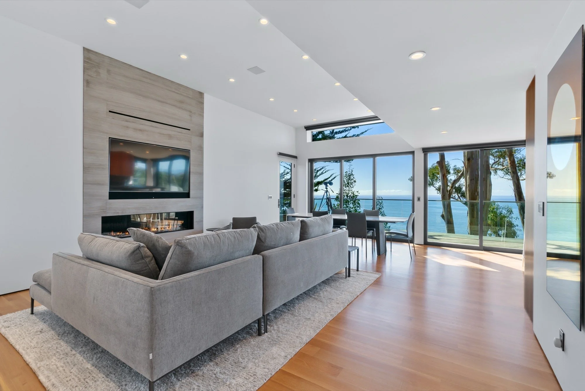 Modern living room with a gray sofa, a wall-mounted TV above a fireplace, large glass windows overlooking water and trees, wooden floors, and minimal decor.