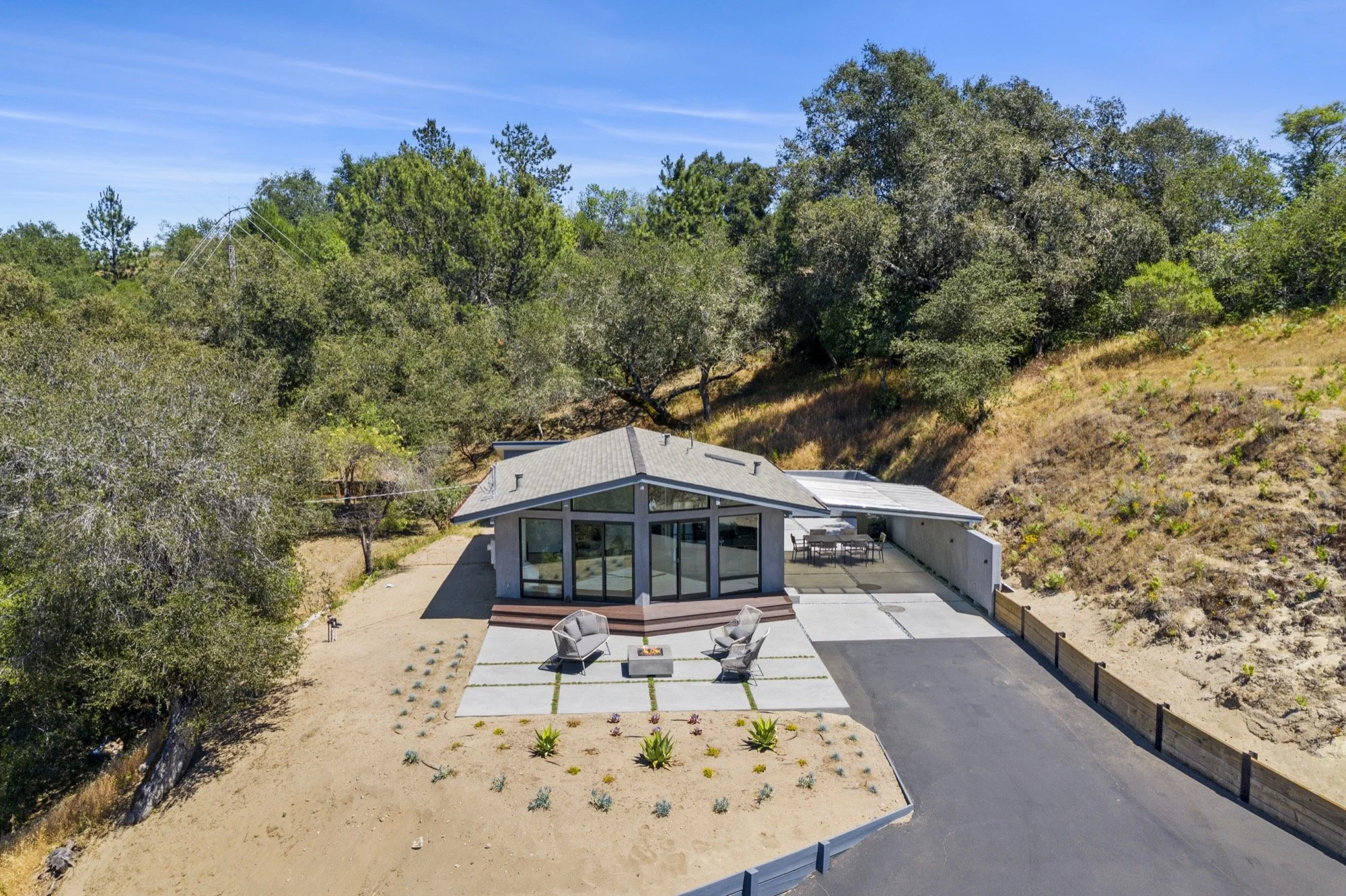 A modern single-story house with large glass windows and a patio, surrounded by trees and dry shrubbery on a hillside, under a clear blue sky.
