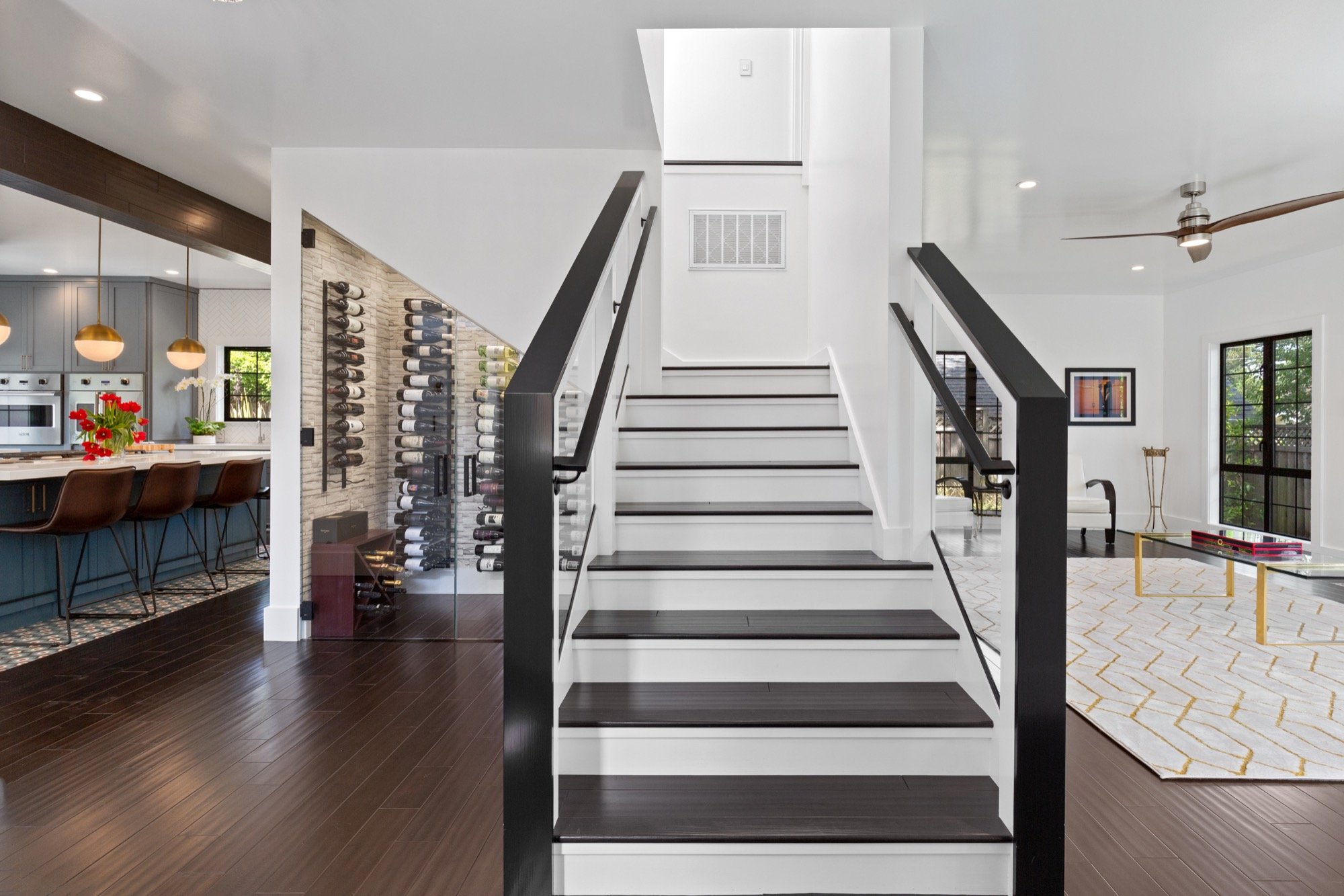 Interior view of a modern house with a staircase in the center, dark wood flooring, a wine wall on the left, and a living room on the right featuring large windows, a white area rug, and contemporary furniture.