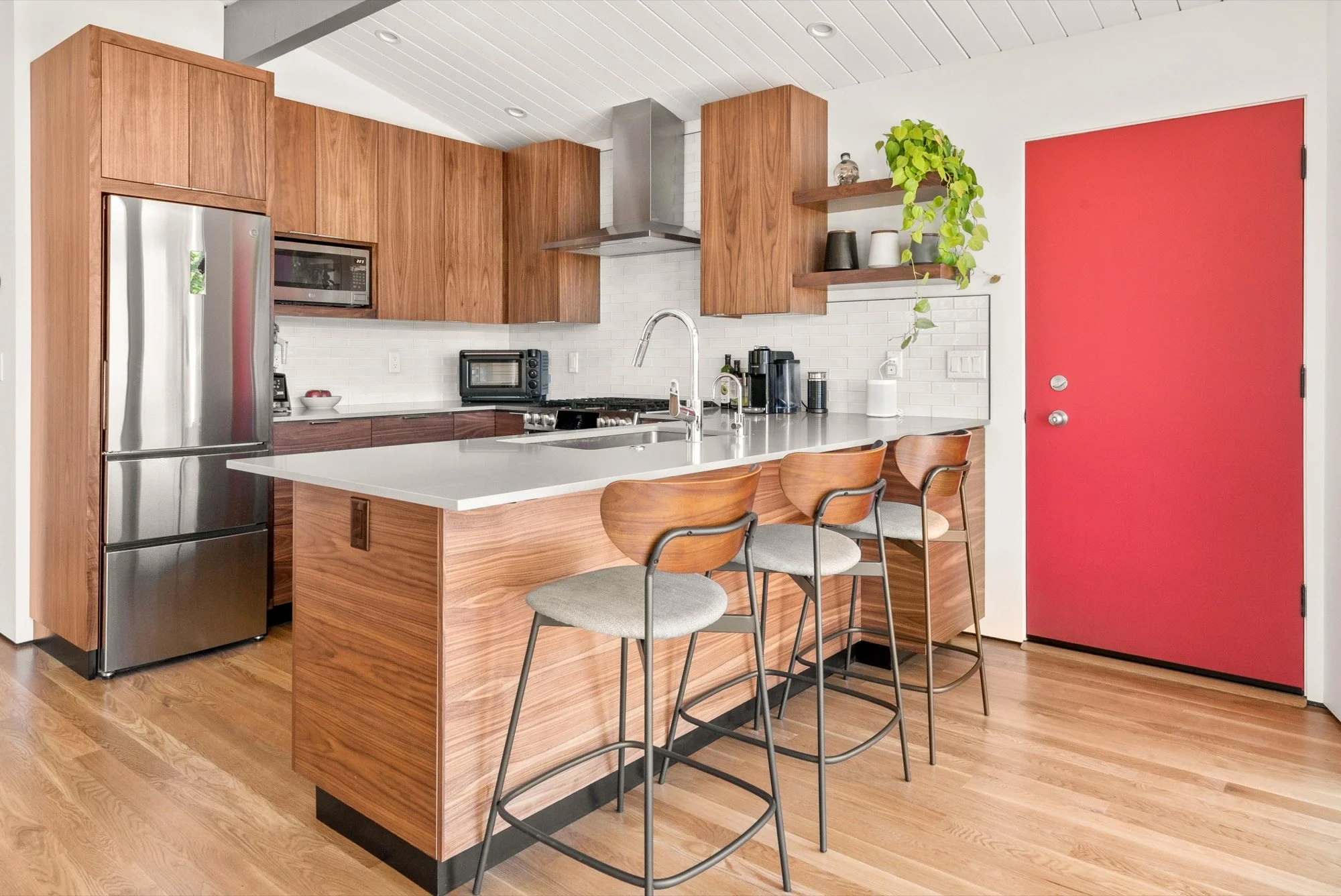 Modern kitchen with wooden cabinets, white countertops, a stainless steel refrigerator, and a red door. There are four barstools with wooden seats and gray cushions at the island, and open shelves with plants and kitchen items.