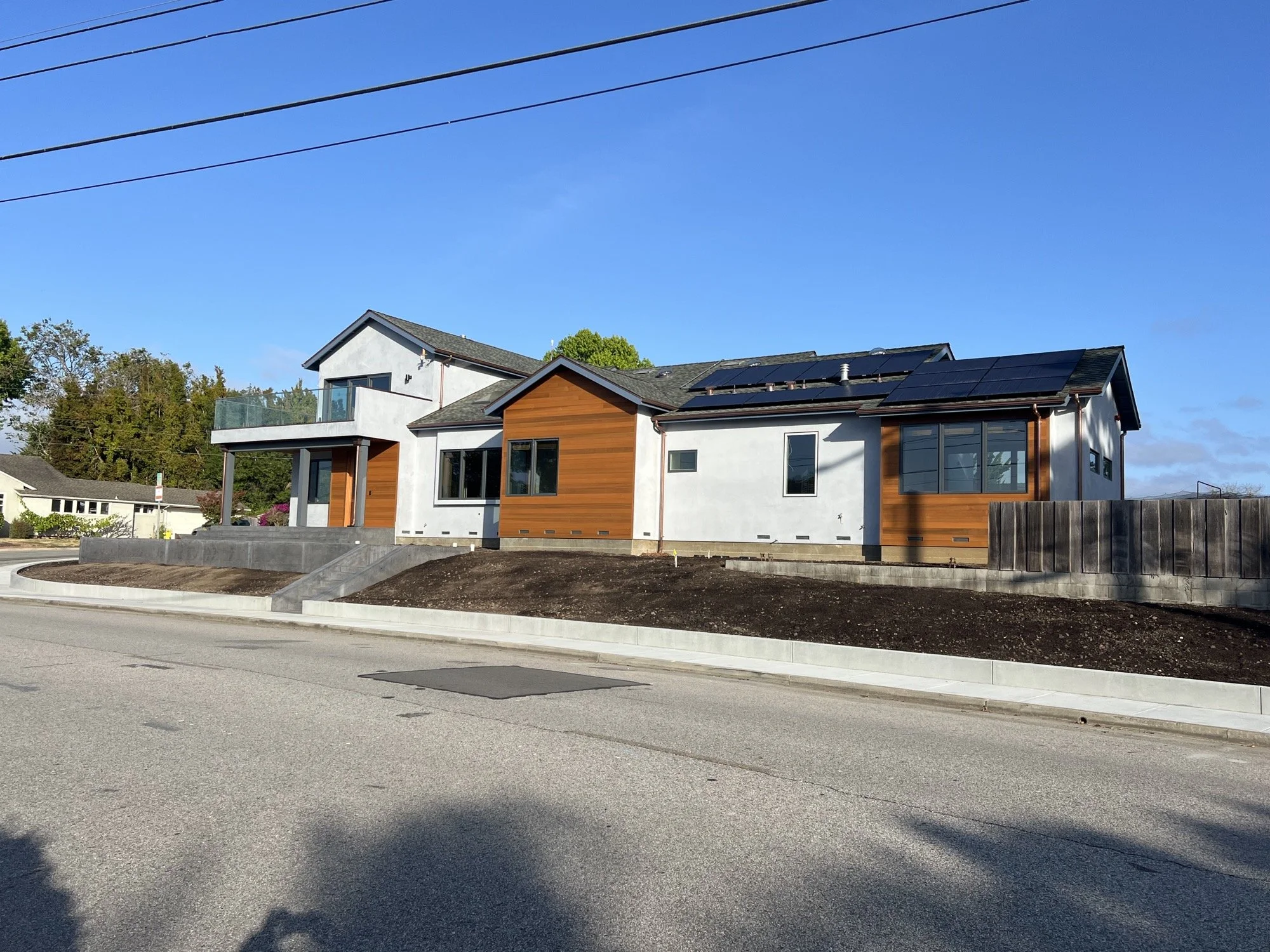 Modern two-story house with white exterior walls, wooden accents, a small front porch, and solar panels on the roof. The house has a sloped driveway and is situated in a suburban neighborhood under a clear blue sky.