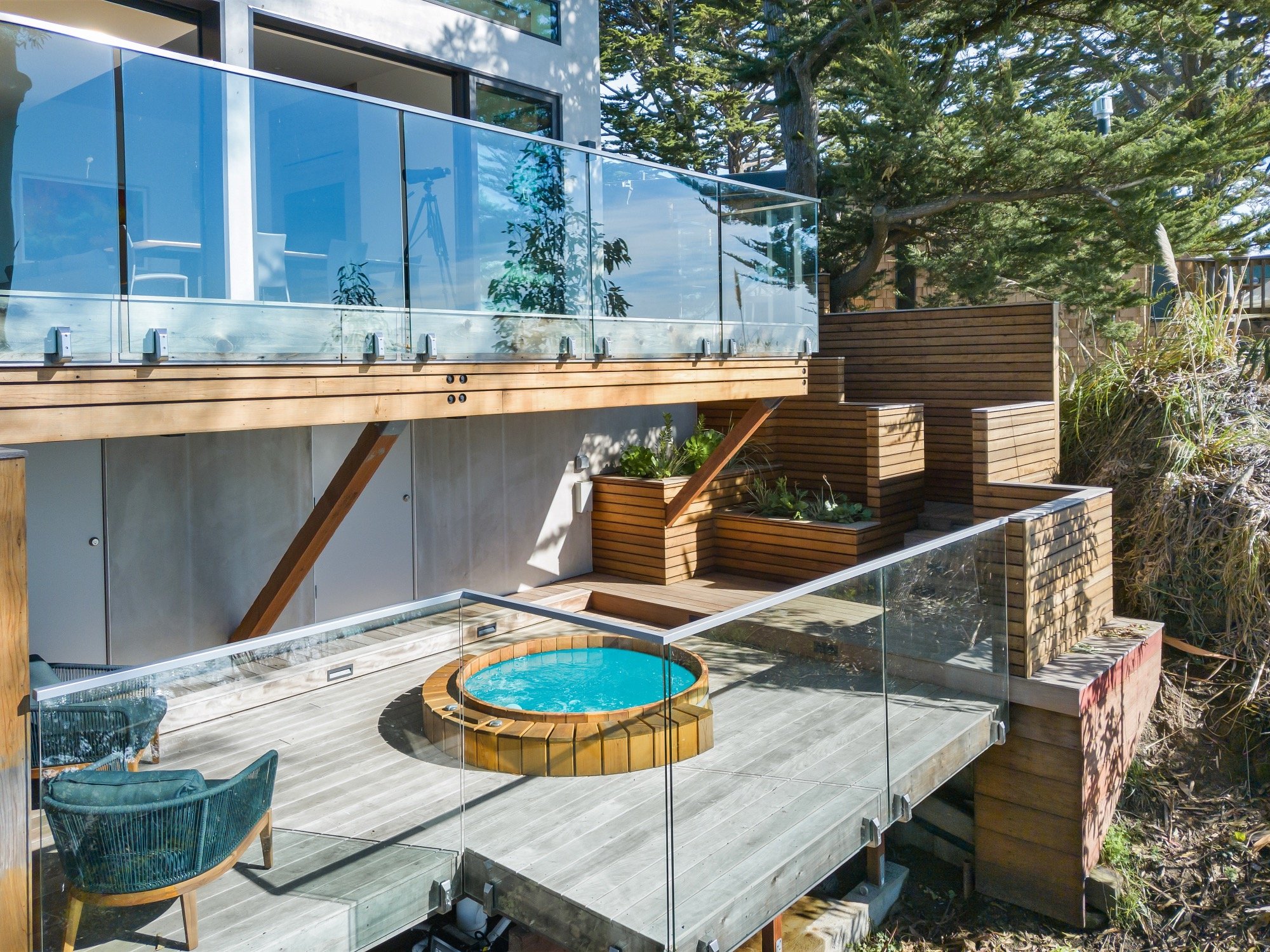 Modern outdoor deck with wooden flooring, glass railing, two chairs around a small round hot tub, with stairs leading to upper level, and outdoor plants, trees, and houses in the background.