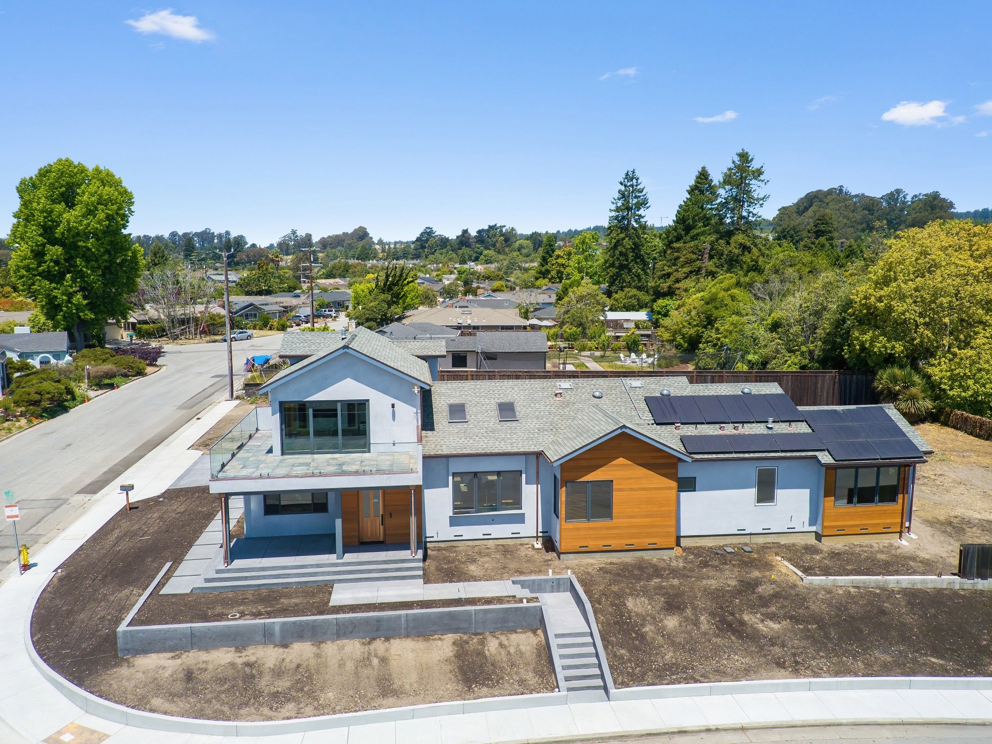 A modern two-story house with blue and wooden exterior, solar panels on the roof, large windows, a balcony, a front porch, an unfinished yard, and a curved concrete driveway in a suburban neighborhood.