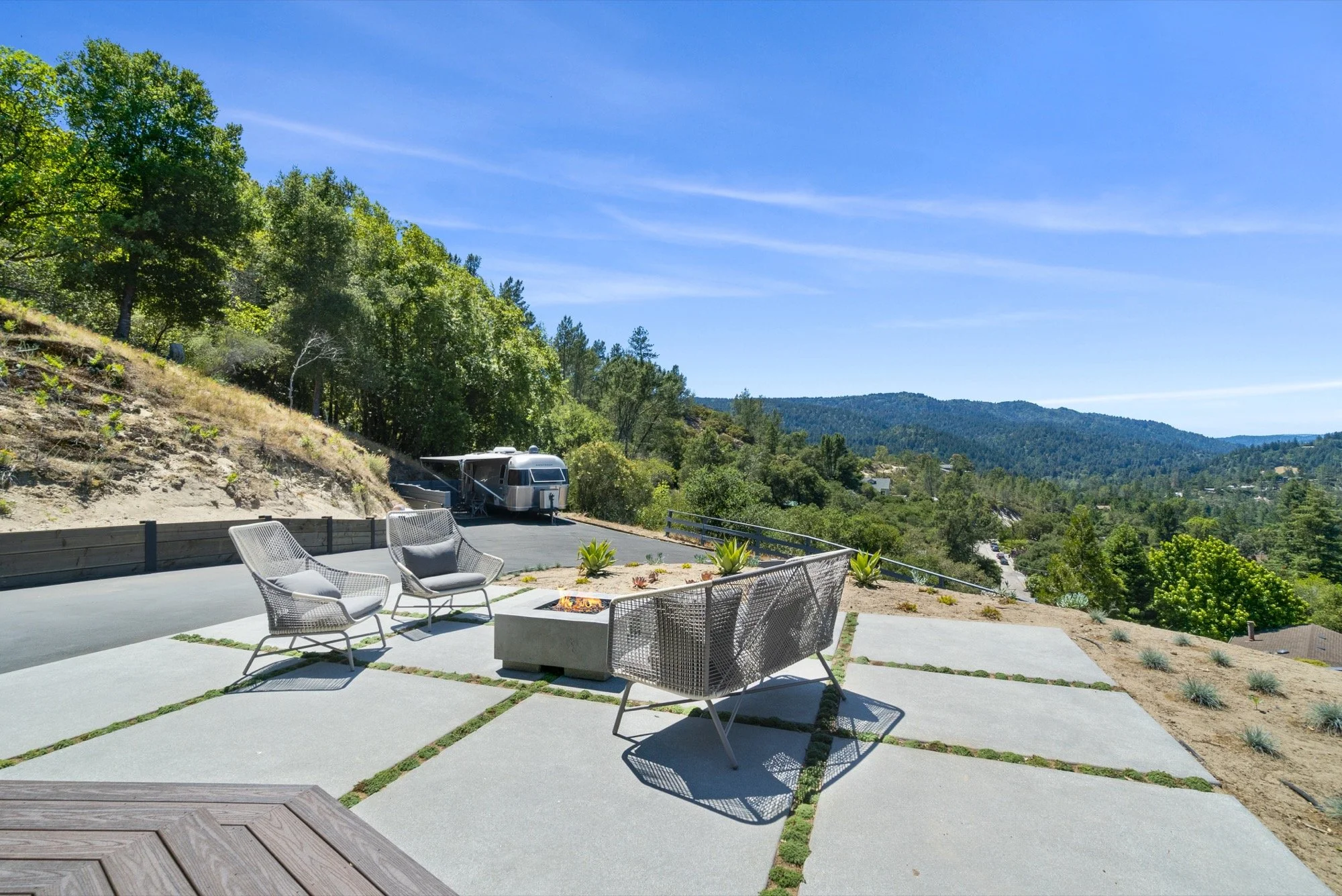 Outdoor patio with four modern chairs around a fire feature, overlooking a mountainous landscape with trees and blue sky.