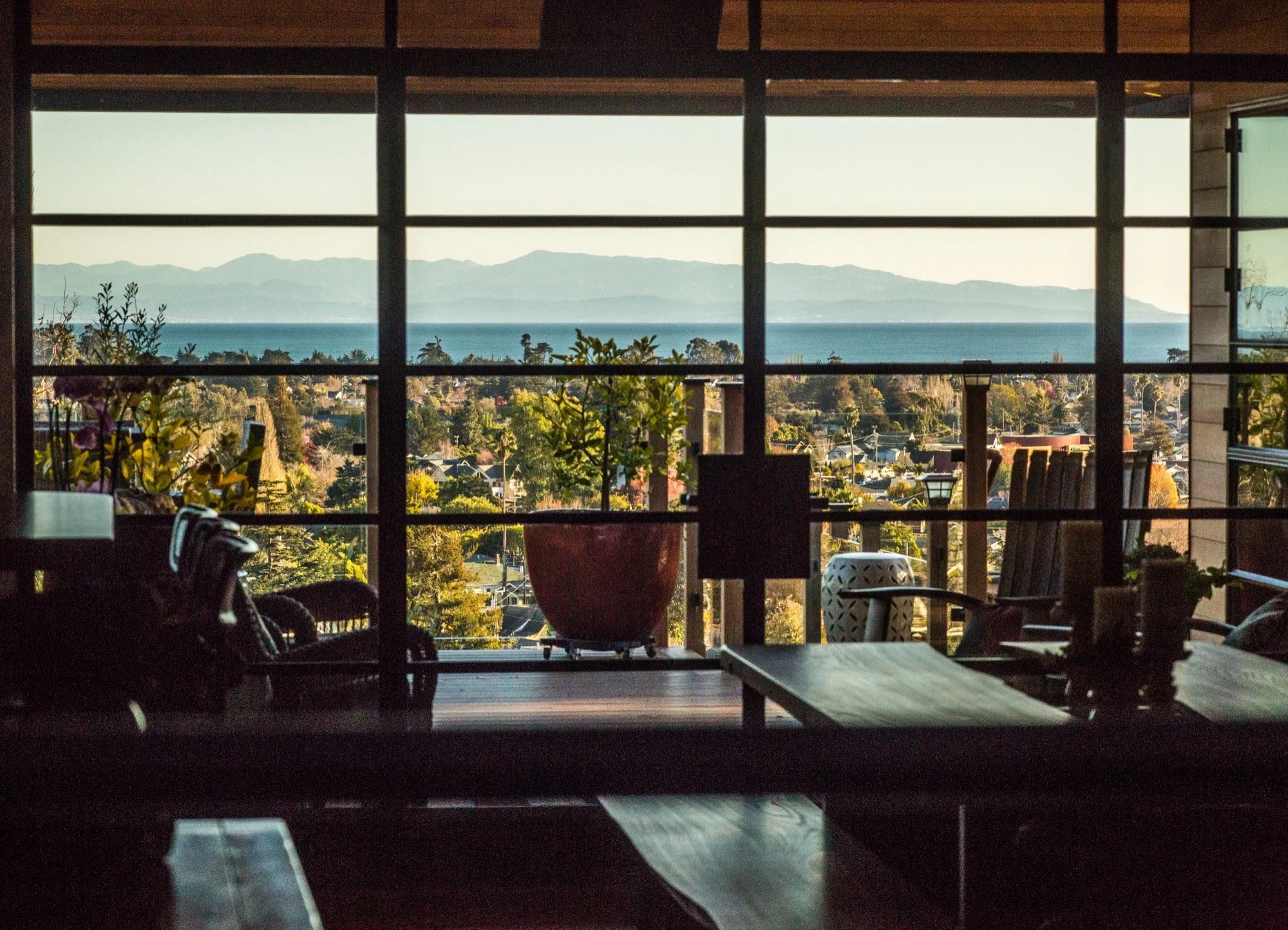 Room with large window overlooking a coastal landscape with mountains, ocean, trees, and houses.