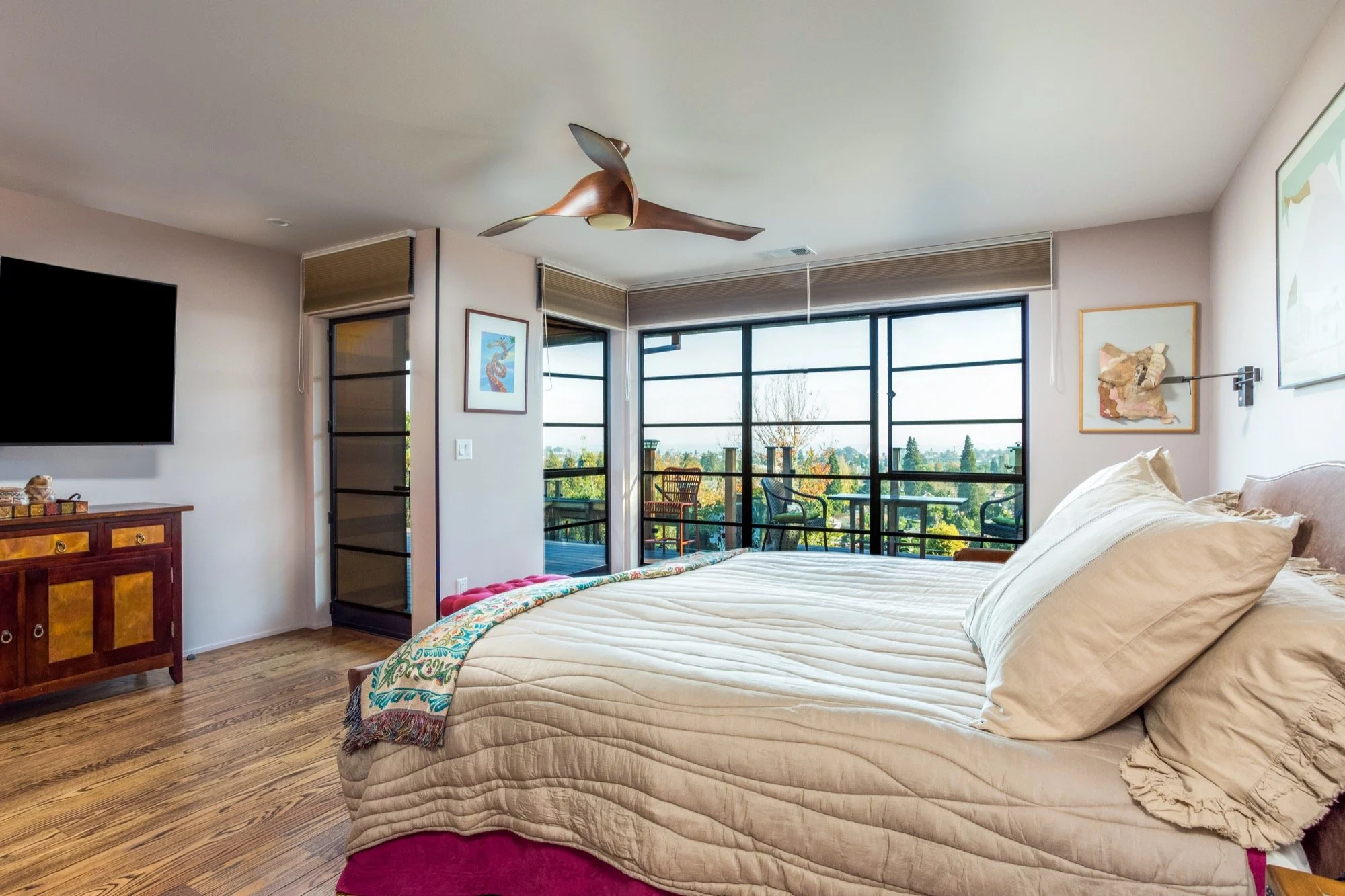 Bedroom with large window overlooking trees, with beige bedding, pillows, and artwork on the walls.
