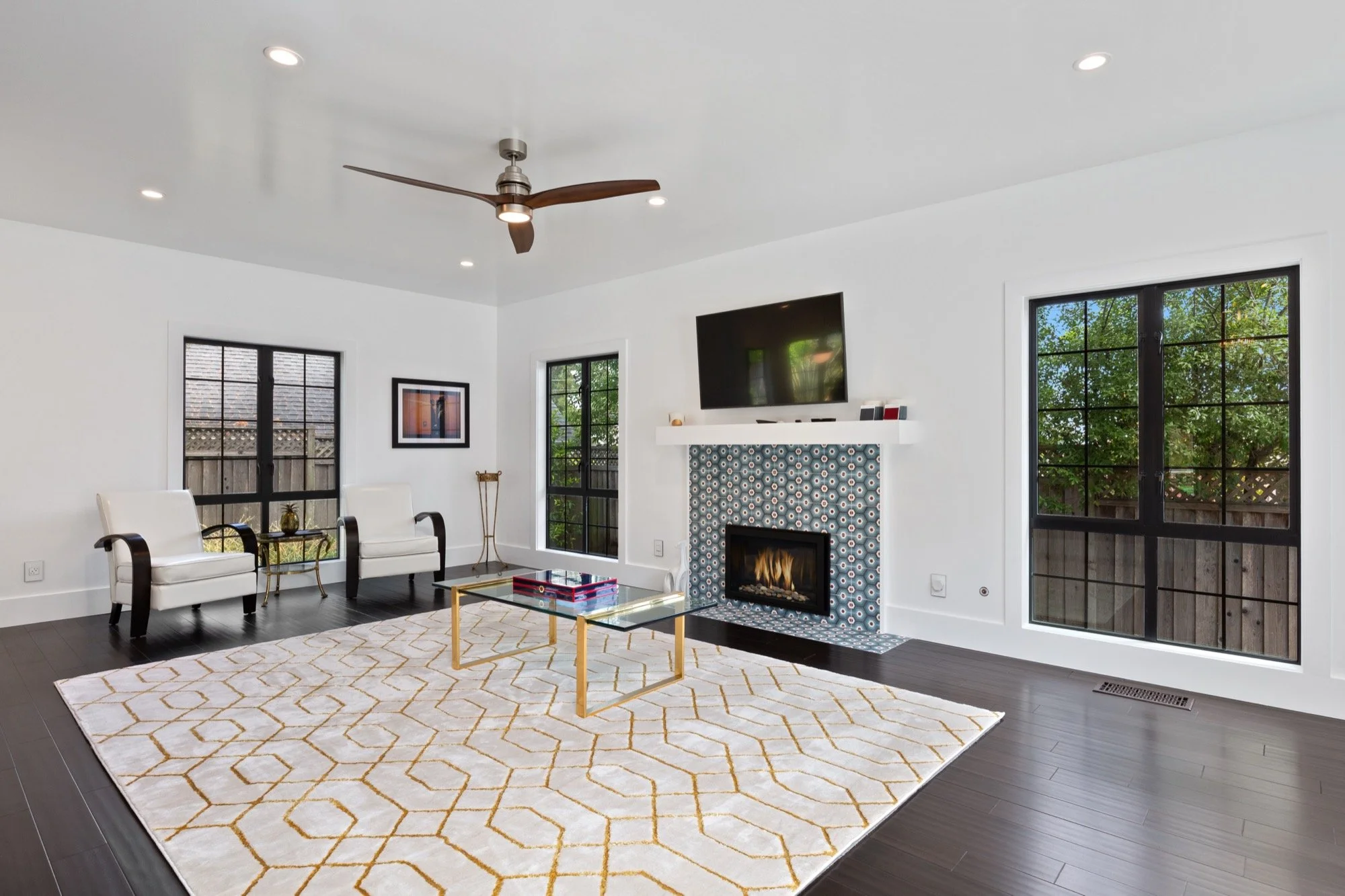 Living room with white walls, dark hardwood floors, large windows showing outdoor trees, a wall-mounted flat-screen TV, a fireplace with decorative tile surrounding, a white rug with a gold geometric pattern, two white armchairs, a glass coffee table