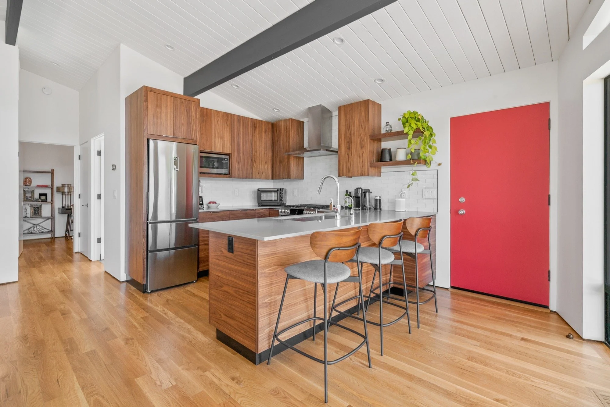 Modern kitchen with wooden cabinets, stainless steel fridge, white countertops, bar stools, and a red door.