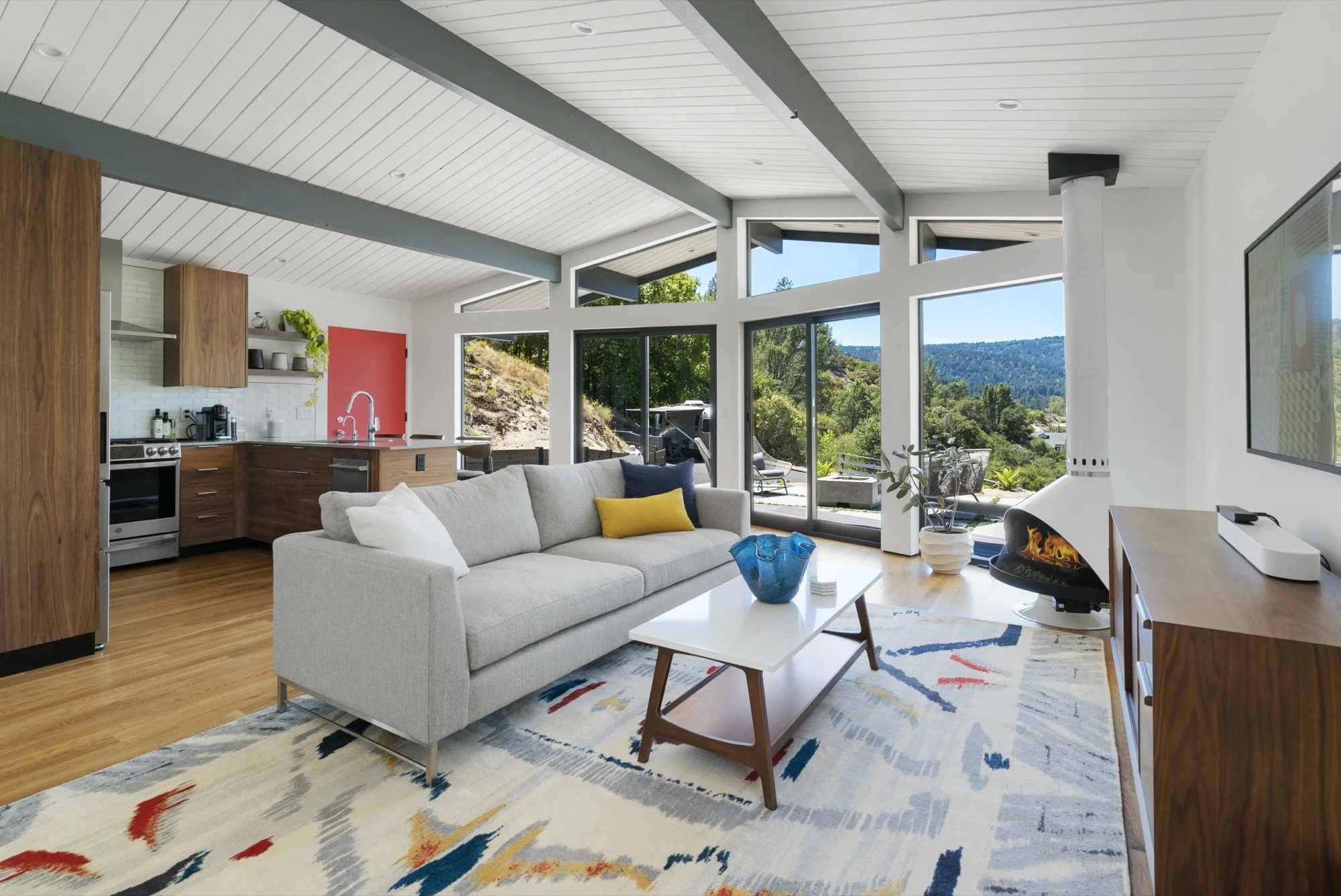 Living room with a gray sofa, colorful pillows, a coffee table with a blue bowl, a white fireplace, large windows showing a scenic outdoor view, a flat-screen TV, and a modern kitchen in the background.