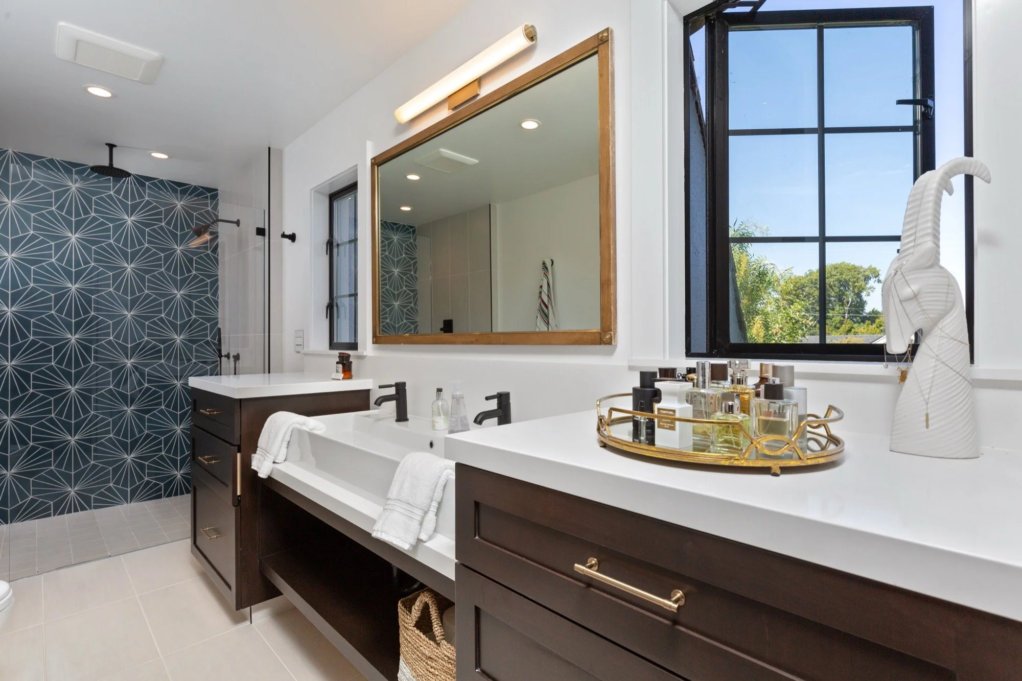 Modern bathroom with double vanity, large mirror, white countertop, black fixtures, decorative drawer pulls, and a tray of toiletries. Features a window with a scenic view and a walk-in shower with blue geometric tile wall.