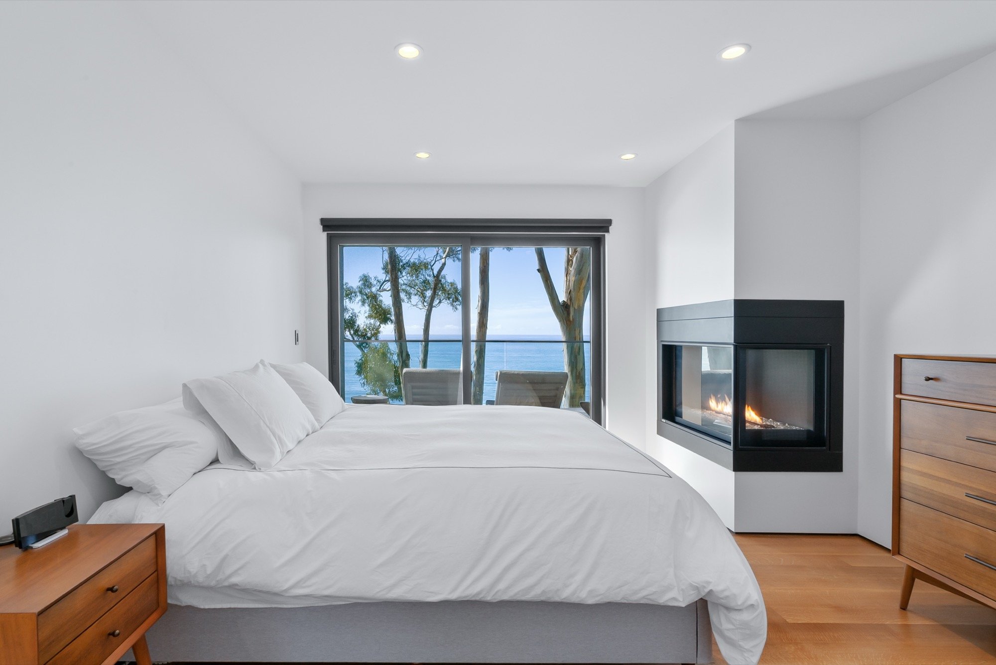 A modern bedroom with white walls, a large bed with white bedding, a wooden nightstand, a fireplace, and a sliding glass door leading to a balcony with outdoor chairs overlooking trees and the ocean.