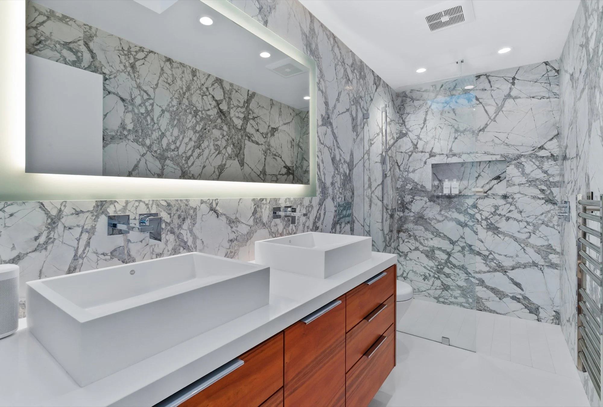 Modern bathroom with marble walls and a double vanity with rectangular vessel sinks, a large mirror with backlit lighting, and a walk-in shower with built-in niche and glass door.