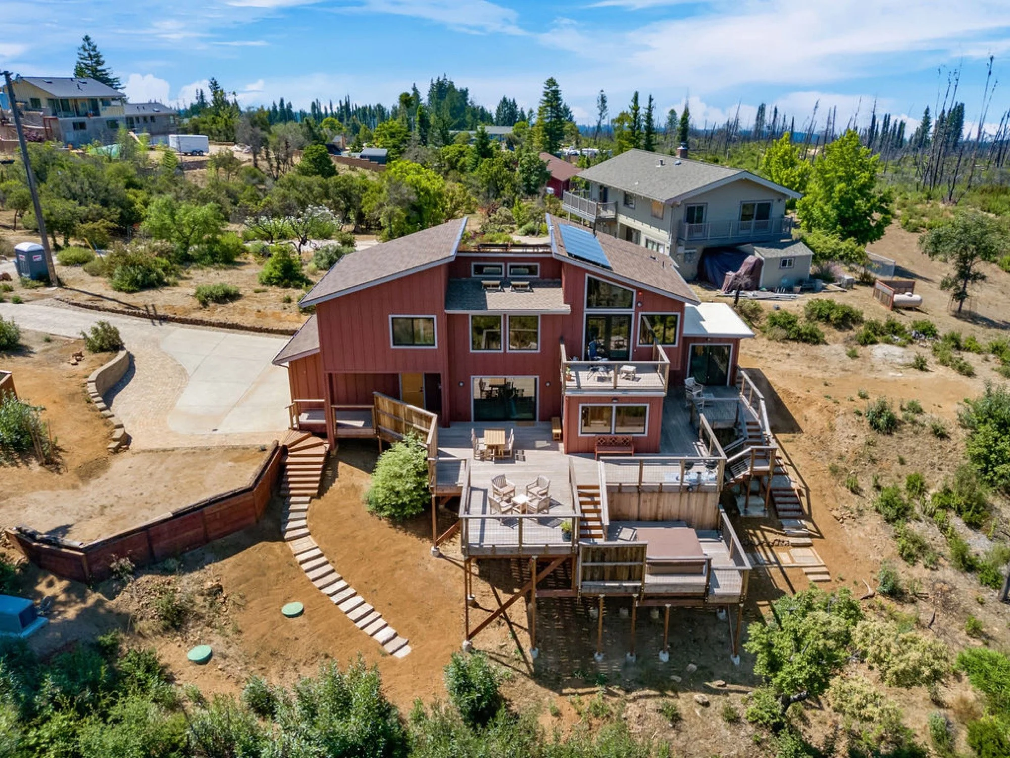 Aerial view of a red house with multiple decks, surrounded by a garden and neighboring houses, in a semi-rural area with trees and open spaces.