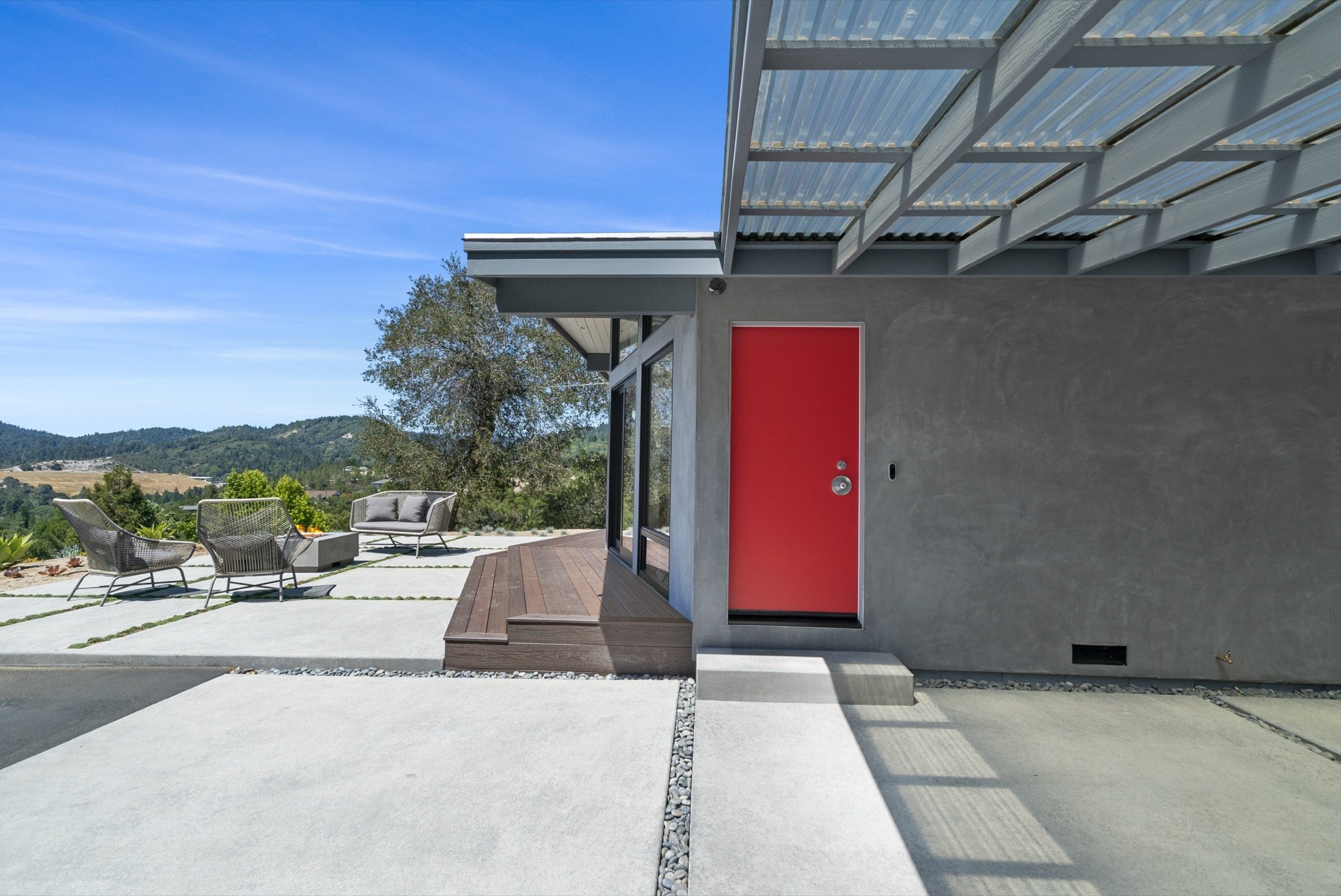 Modern house exterior with a bright red door, concrete and wooden steps, glass walls, metal roof, outdoor seating area, and hill landscape in the background