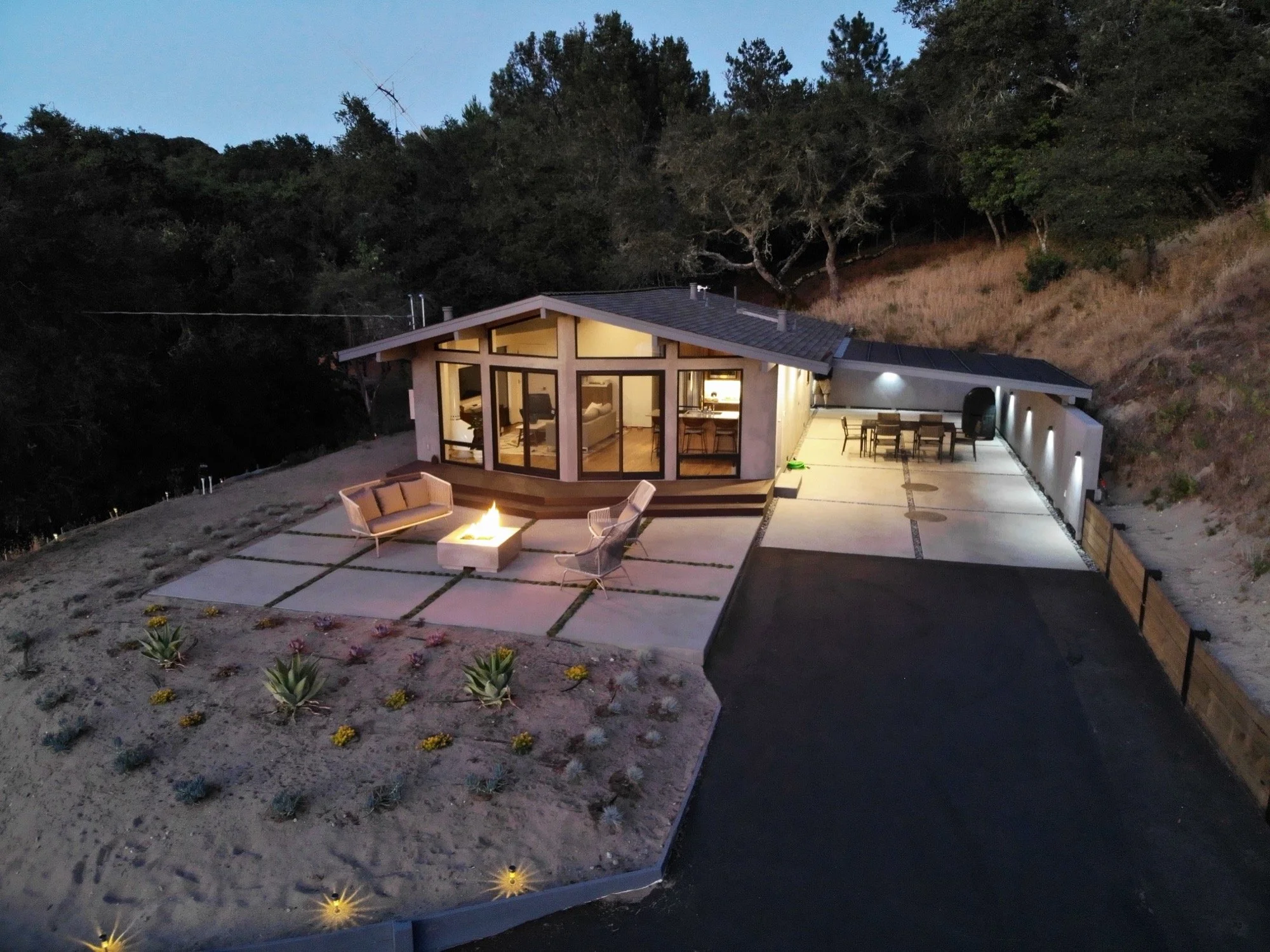 A modern house with large glass windows, illuminated at dusk, situated on a hillside. The house features an outdoor patio with seating, a fire pit, and surrounding landscaping with plants and pathway lighting.