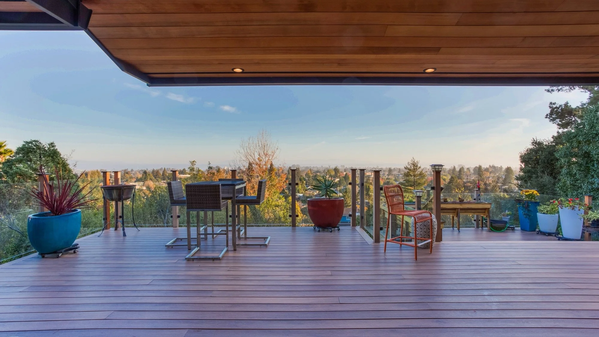 Empty wooden balcony with potted plants, outdoor furniture, and a view of trees and sky in the distance.