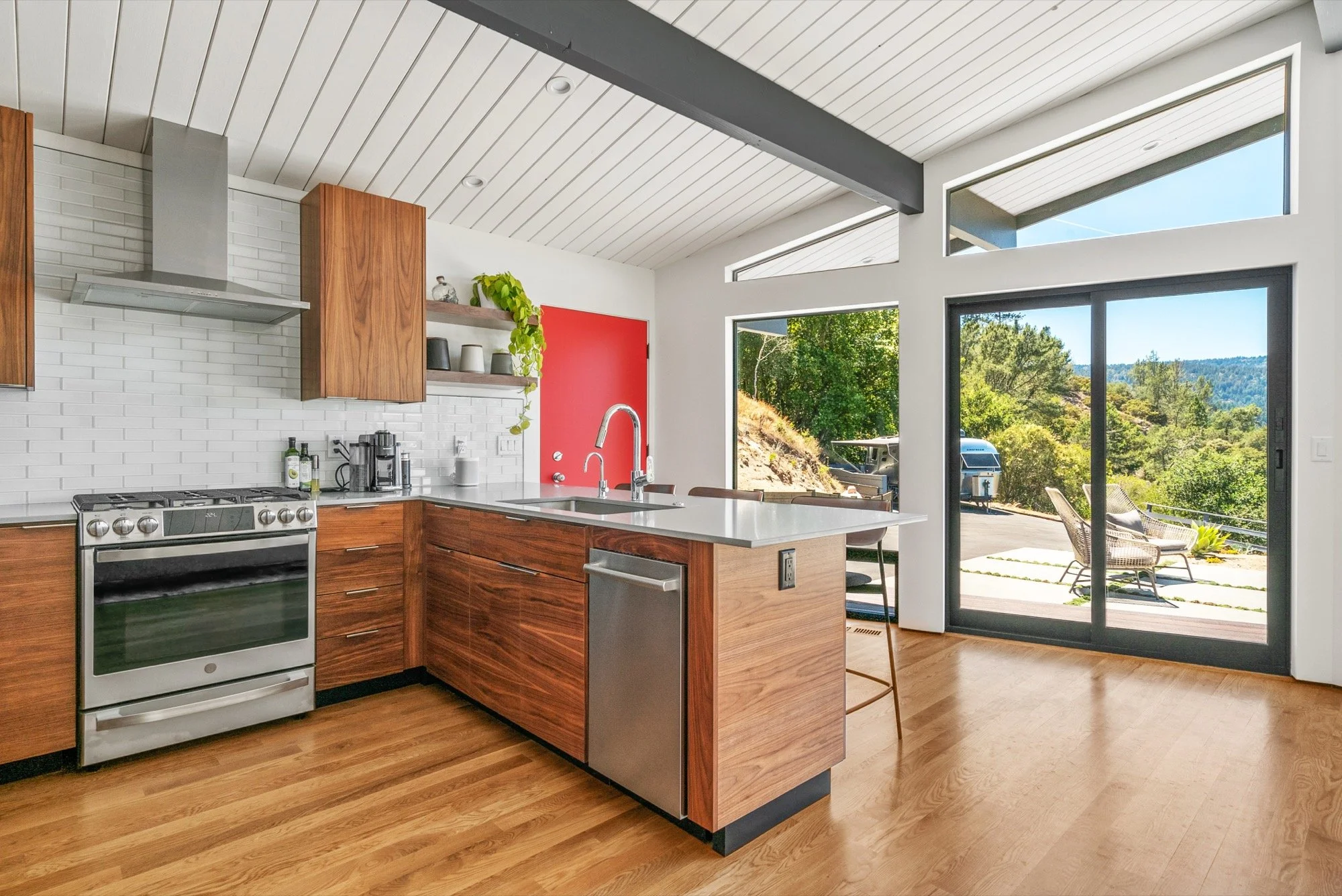 Modern kitchen with wooden cabinets, white countertops, stainless steel appliances, and large glass sliding doors opening to an outdoor patio with chairs and a scenic view of trees and hills.
