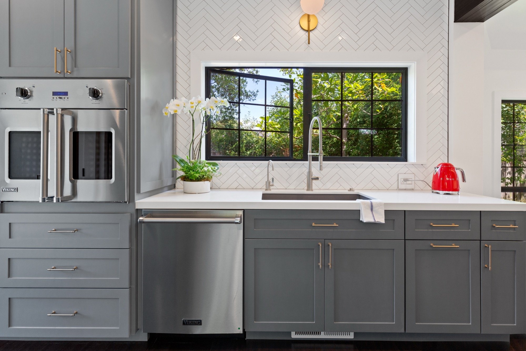 Modern kitchen with gray cabinets, white countertop, stainless steel appliances, black-framed window, orchids, red kettle, and greenery outside.