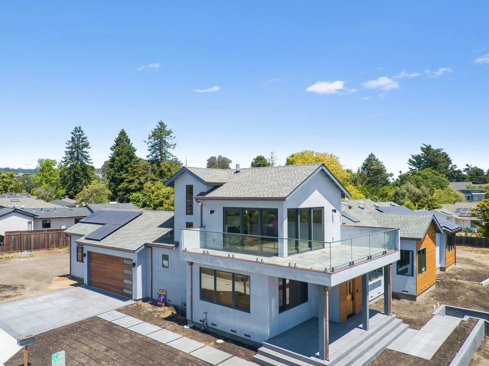 A modern two-story house with a glass balcony, large windows, and a mix of white stucco and wood siding, situated in a suburban neighborhood with clear skies and trees in the background.