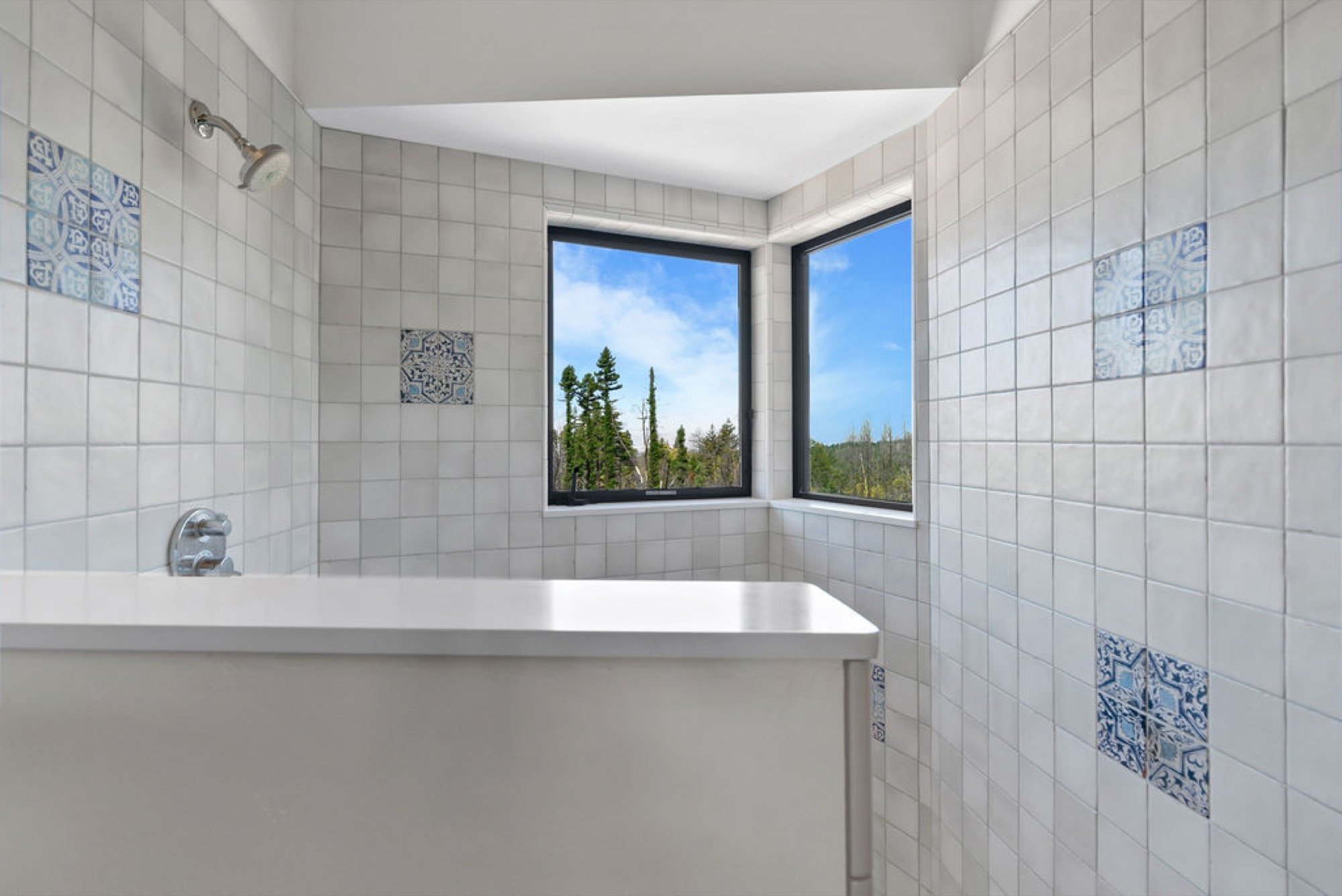 A bathroom with square white tiles and decorative blue patterned tiles, a bathtub, and two large windows showing a view of trees and a blue sky.