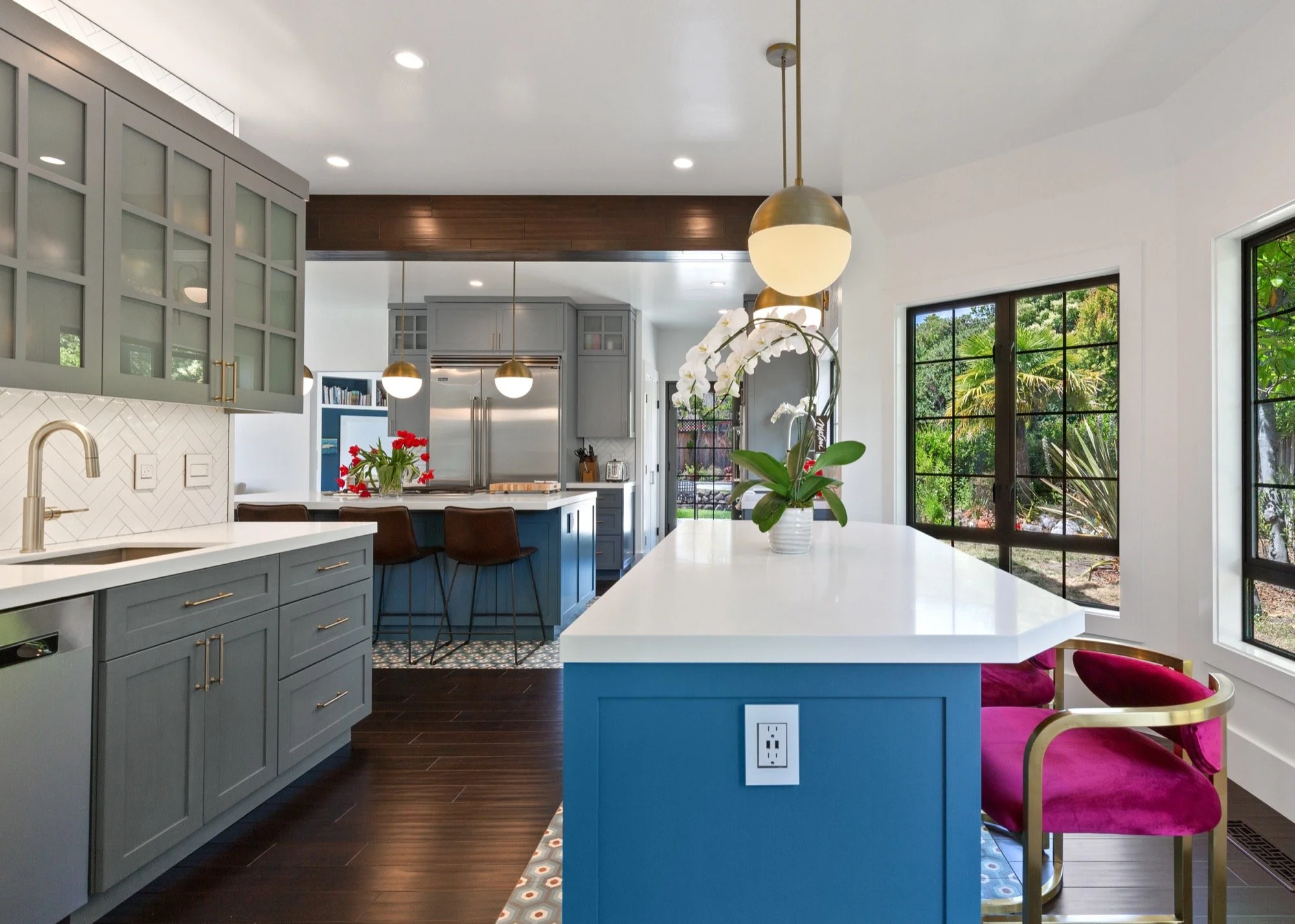 Modern kitchen with blue cabinetry, white countertops, large windows, and pendant lighting, featuring a potted orchid on a breakfast bar and a glass door leading outside.