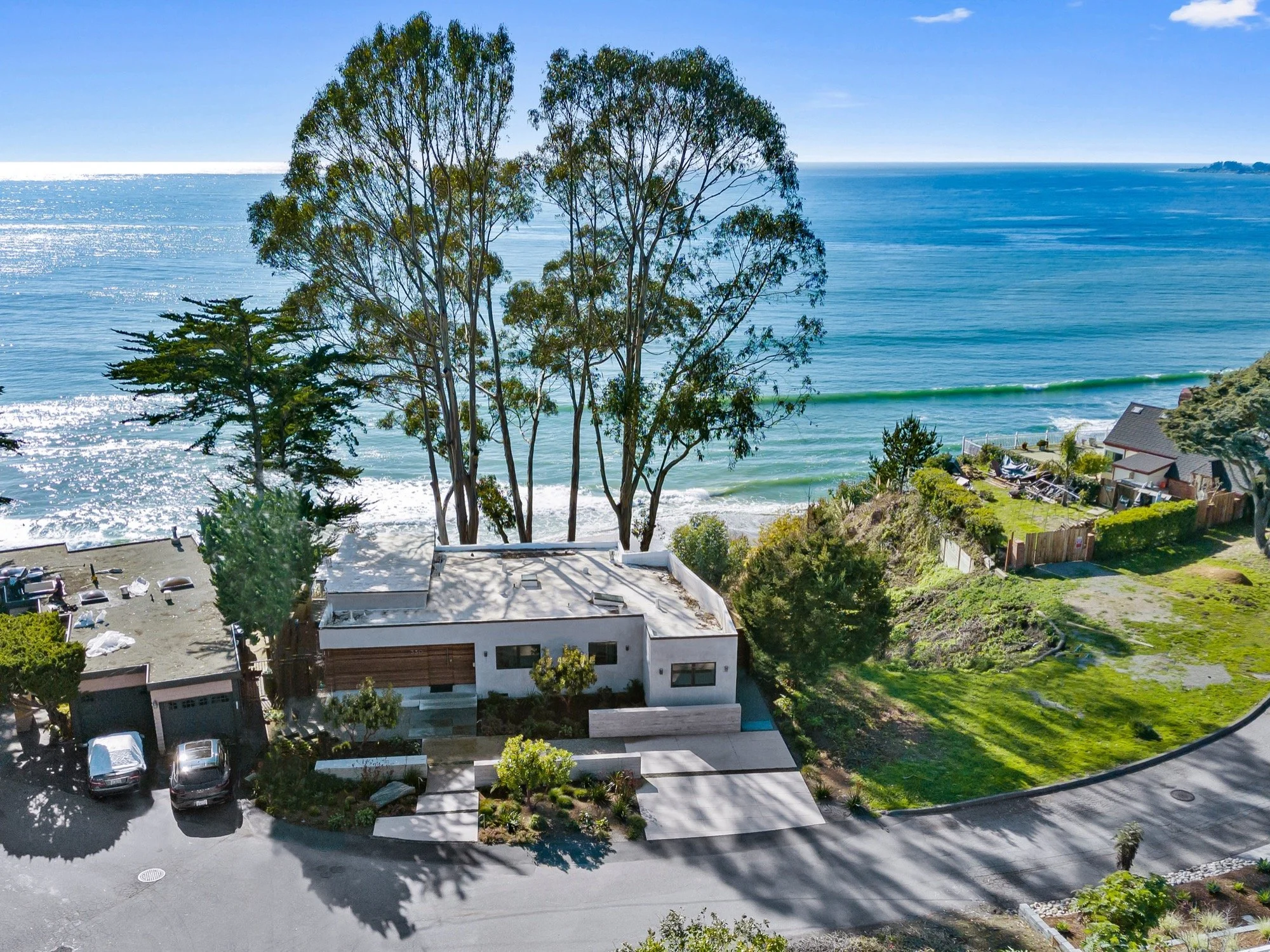 Aerial view of a modern house with a flat roof, surrounded by trees and landscaped yard, overlooking the ocean with waves and a clear sky.
