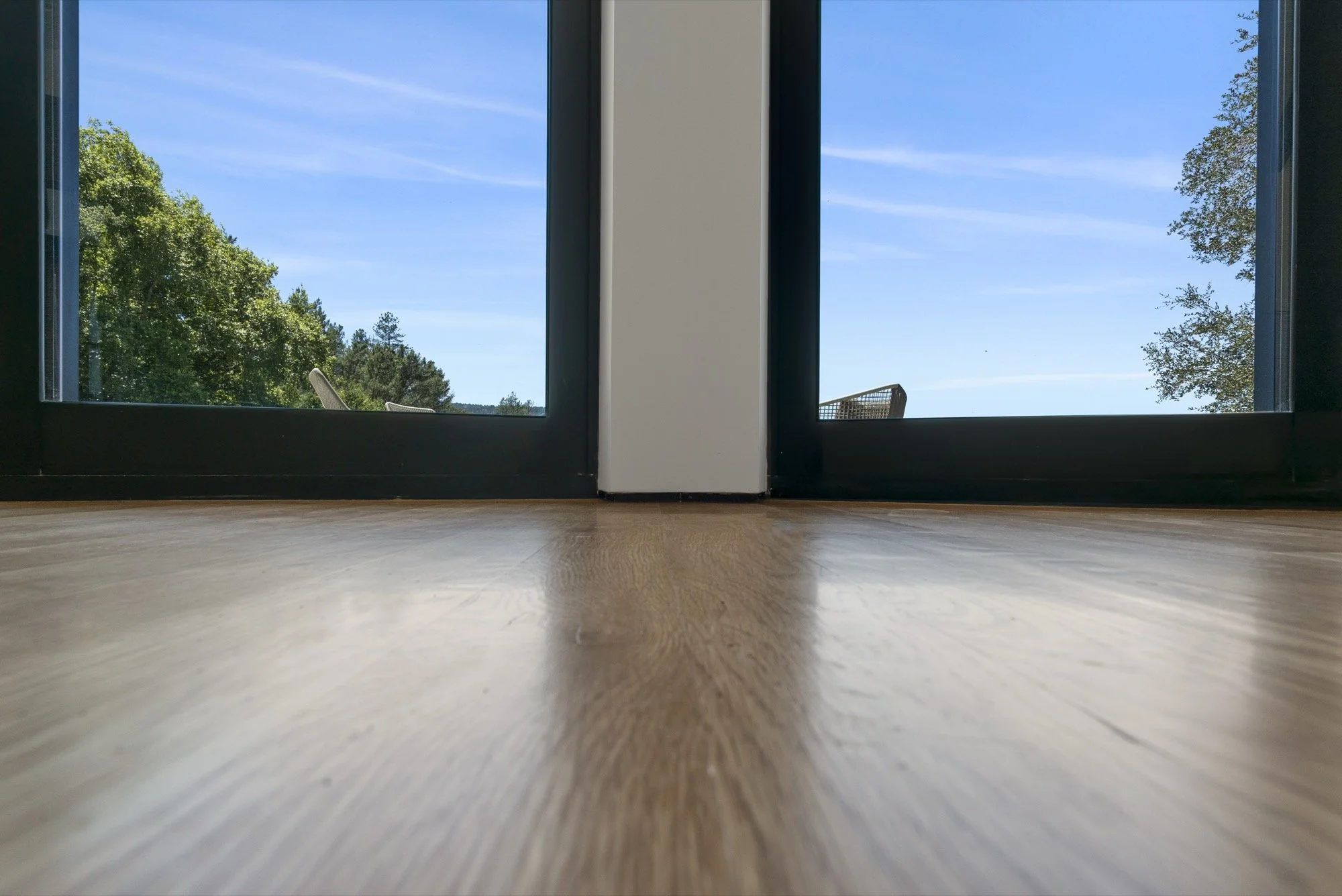 Low-angle view of glass sliding doors with a wooden floor inside, showing a clear blue sky and green trees outside.