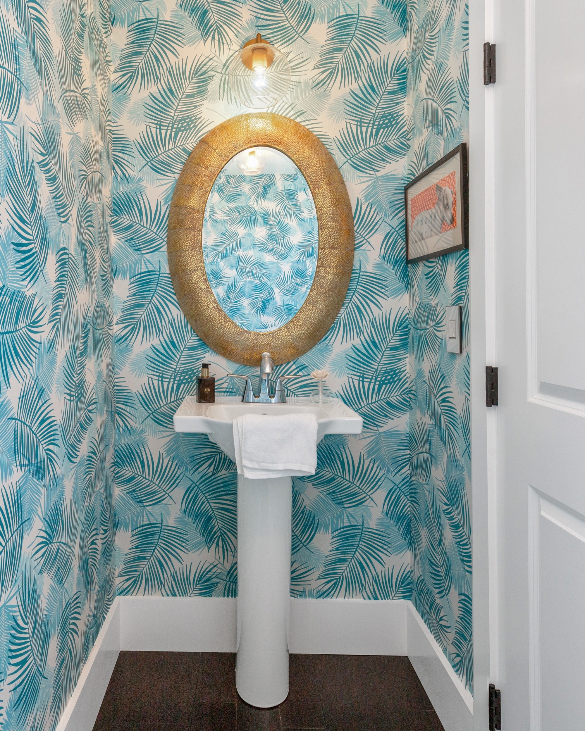 Small bathroom with tropical leaf wallpaper, oval mirror with gold frame, white pedestal sink with a white towel, soap dispenser, and decorative shell, light wood and glass light fixture, framed artwork of a zebra, and white door.