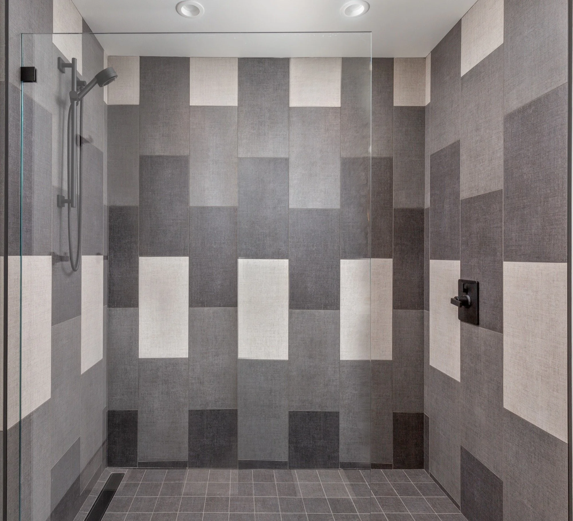 A modern walk-in shower with gray and beige tiled walls and a glass shower door. There is a rainfall showerhead and a handheld showerhead with black fixtures.