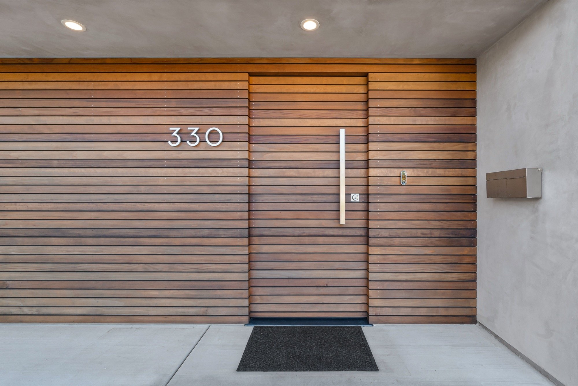 Modern front door with horizontal wooden slats, house number 330 on the left, a vertical handle and doorbell, with a mailbox on the right side wall.