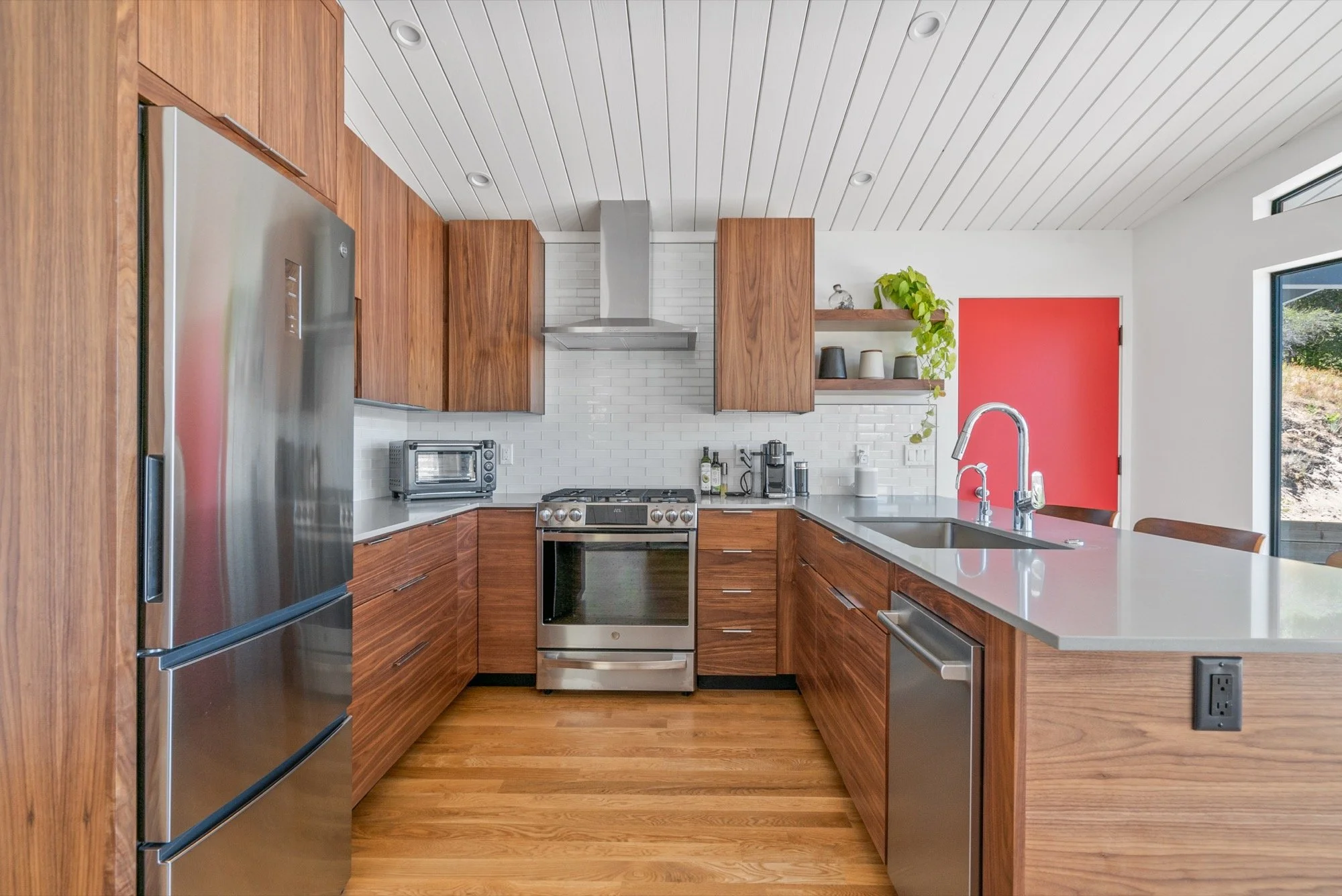 Modern kitchen with wood cabinets, stainless steel refrigerator, oven, and dishwasher, white brick backsplash, gray countertop, and open shelves decorated with plants and dishes.