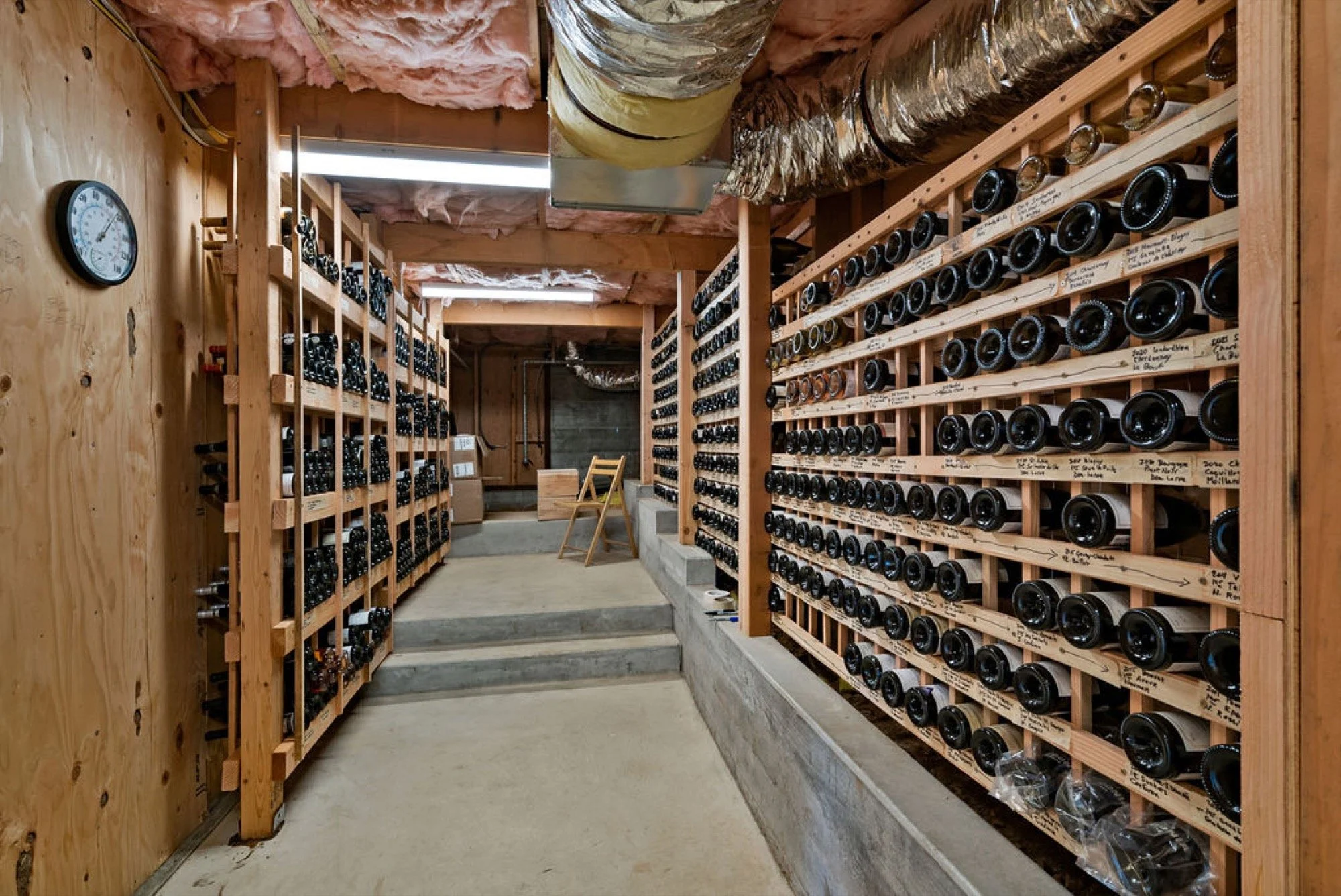 A wine cellar with wooden shelves filled with wine bottles, concrete steps leading to an upper level, and insulation visible on the ceiling.