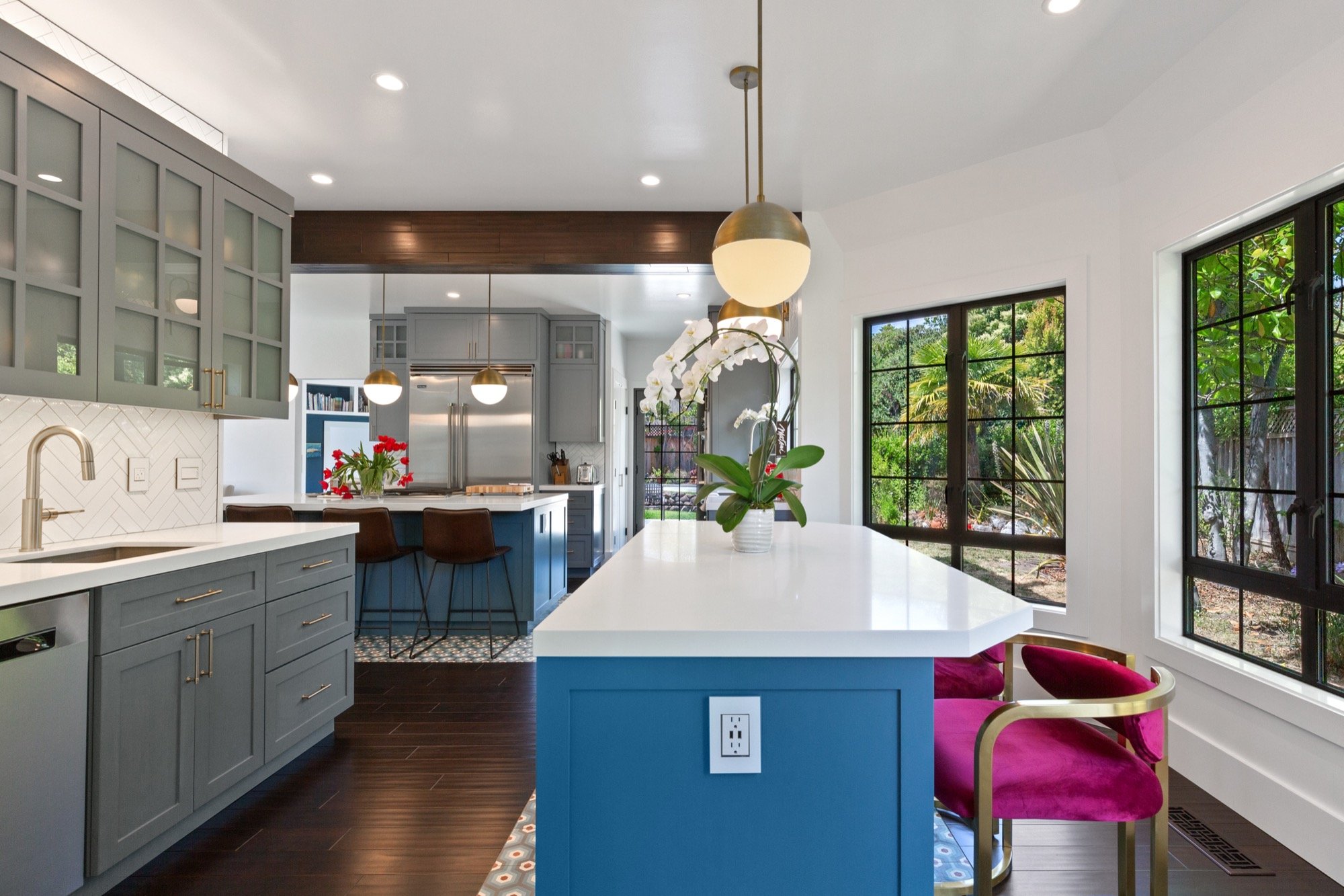 A modern kitchen with a large white island counters, gray cabinets, and black-framed windows showing greenery outside. It has colorful chairs and hanging pendant lights.