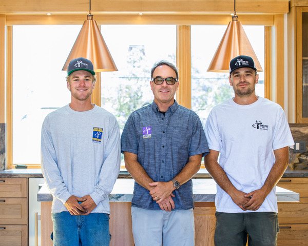 Three men standing inside a kitchen with wooden cabinets and large windows. The man in the middle wears glasses and a blue shirt, while the two men on either side wear baseball caps and casual shirts. All three are smiling.