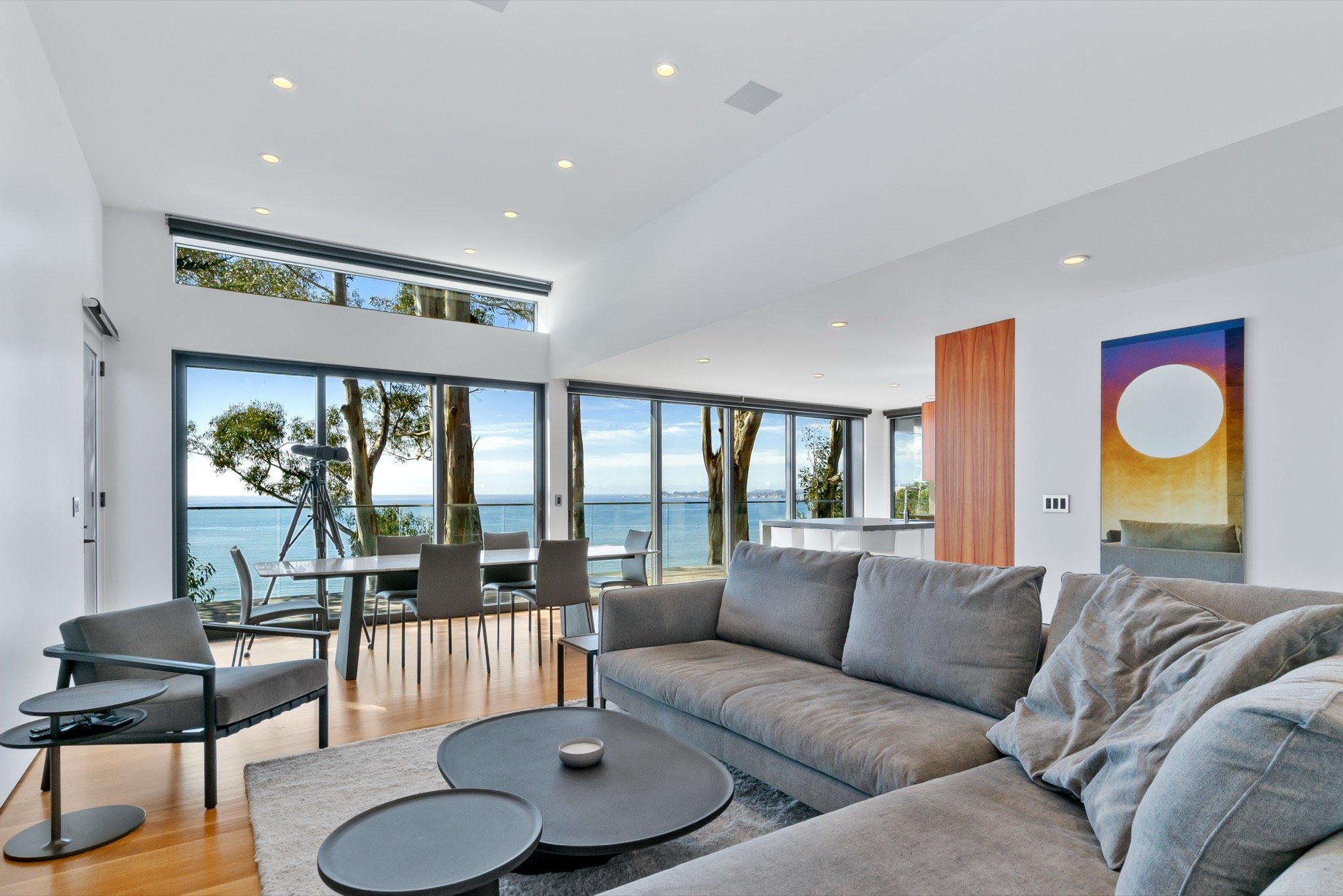 Modern living room with large glass windows overlooking the ocean, with a telescope near the window, gray sofas, a black side table, and a round coffee table, decorated with minimalist art and wood trim.