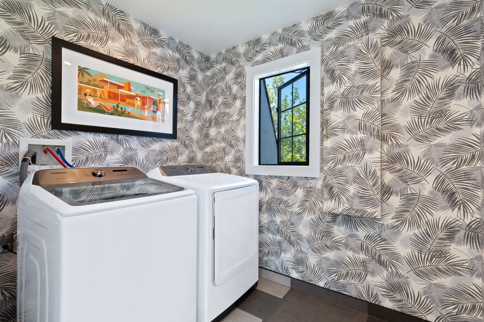 A laundry room with a patterned wallpaper of black and white palm leaves, containing a white top-loading washing machine and a white dryer, with a window showing green trees outside, and a colorful framed art print on the wall.