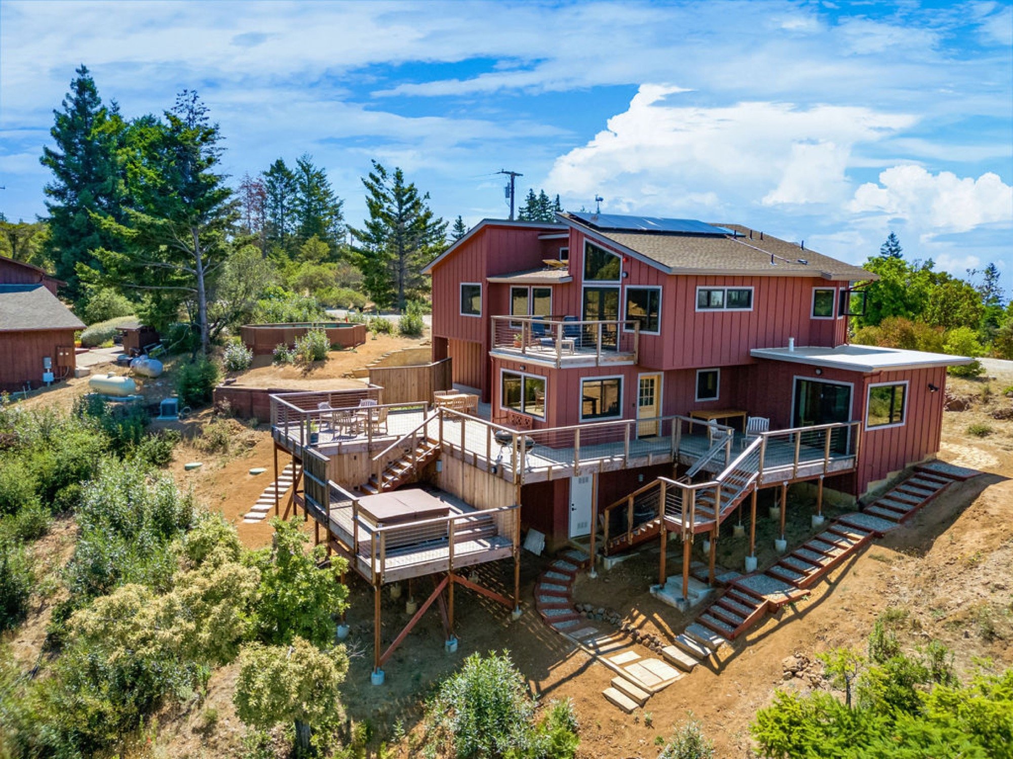 A multi-level modern house with red exterior and large windows, built on a hillside with a spacious wooden deck and stairs, surrounded by trees and greenery, under a partly cloudy sky.