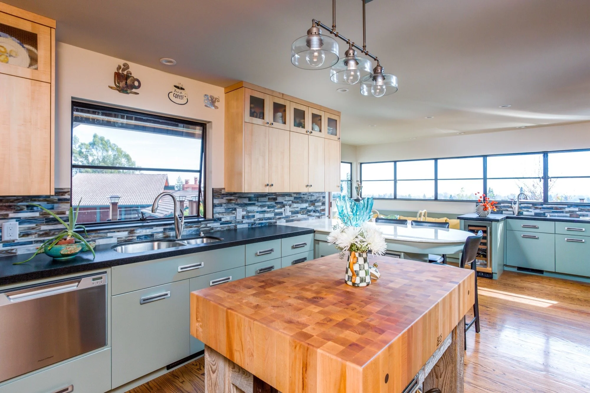 Bright modern kitchen with light green cabinets, black countertops, a wood kitchen island with a checkered vase of white flowers, large windows, and a viewing area in the background.