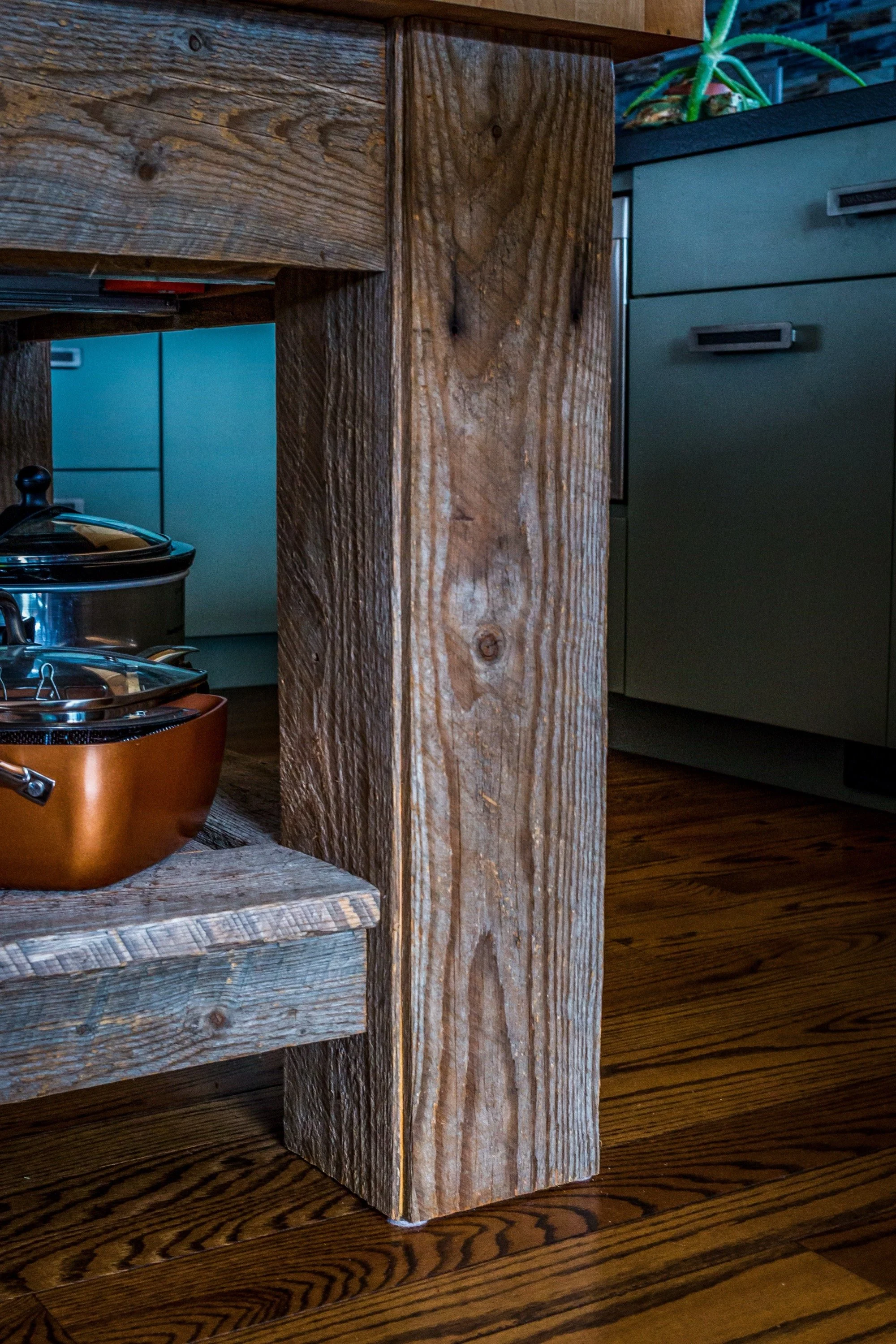 Close-up of a wooden kitchen island leg with metal pots and pans on the shelf underneath, and part of a green cabinet and wooden floor in the background.