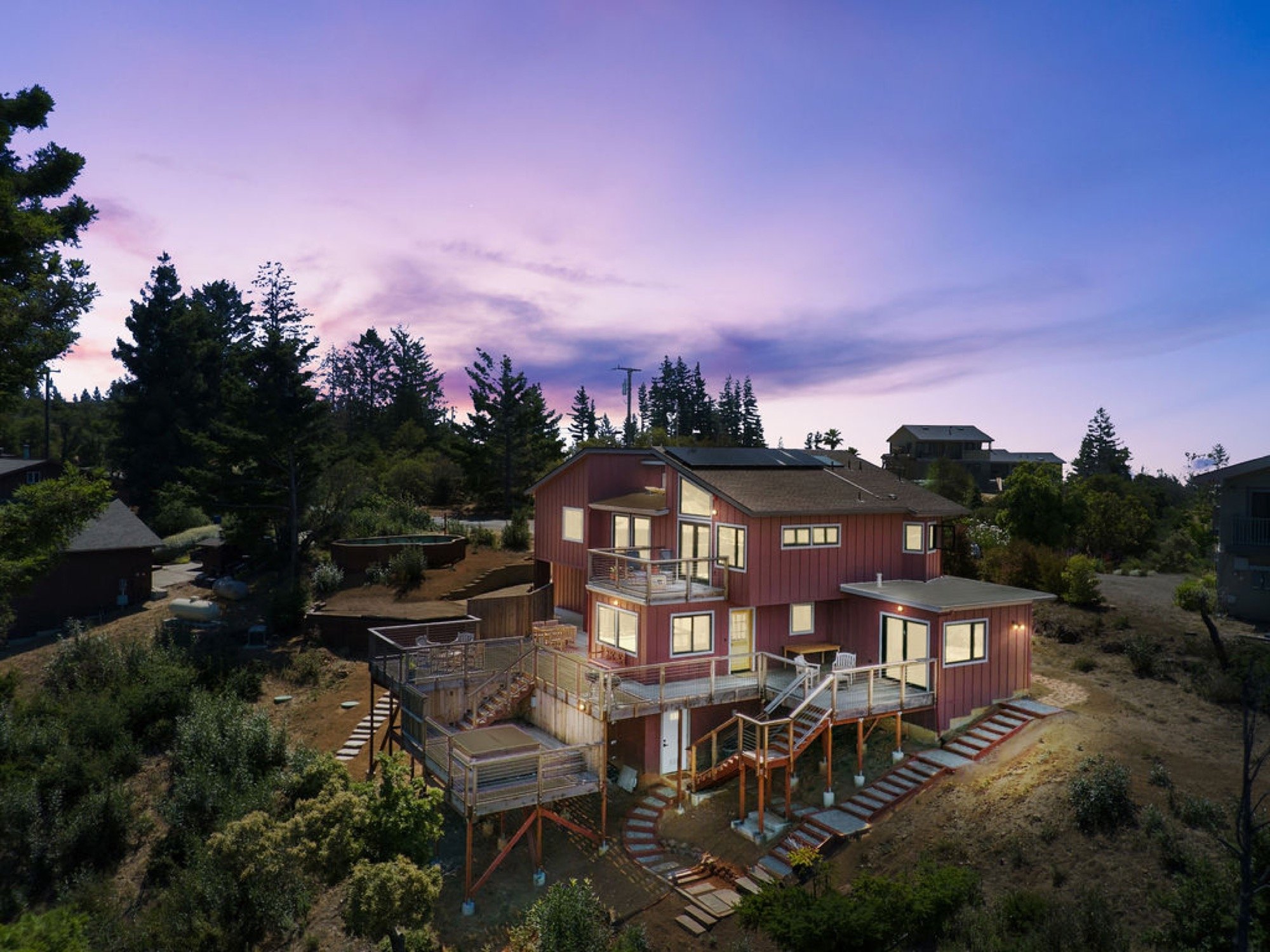 A multi-story house with red exterior walls, large windows, and multiple outdoor decks is situated on a hillside with surrounding trees and vegetation during dusk.