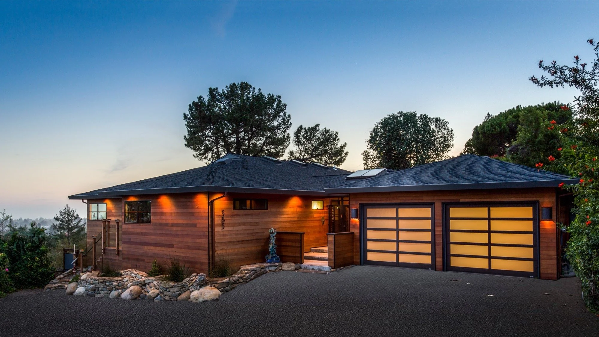 Modern wooden house with a two-car garage, illuminated exterior lighting, surrounded by trees and garden rocks at dusk.