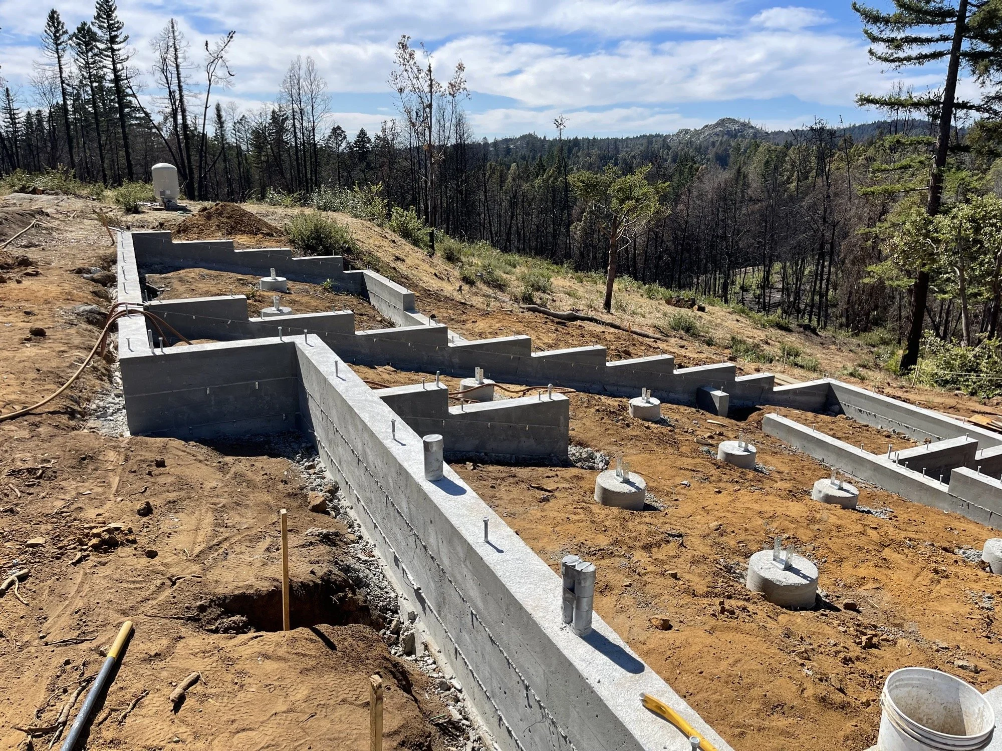 Construction site with concrete foundation and footings on a hillside, surrounded by trees and mountains in the background.