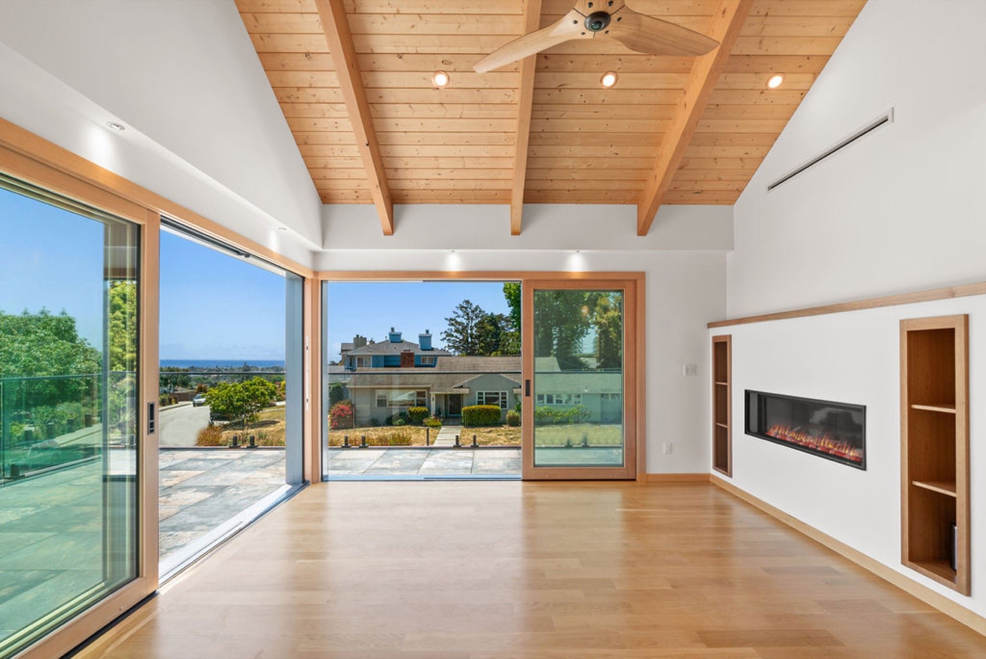 Empty living room with large sliding glass doors, wooden ceiling with exposed beams, and small built-in shelves around an electric fireplace on a white wall.