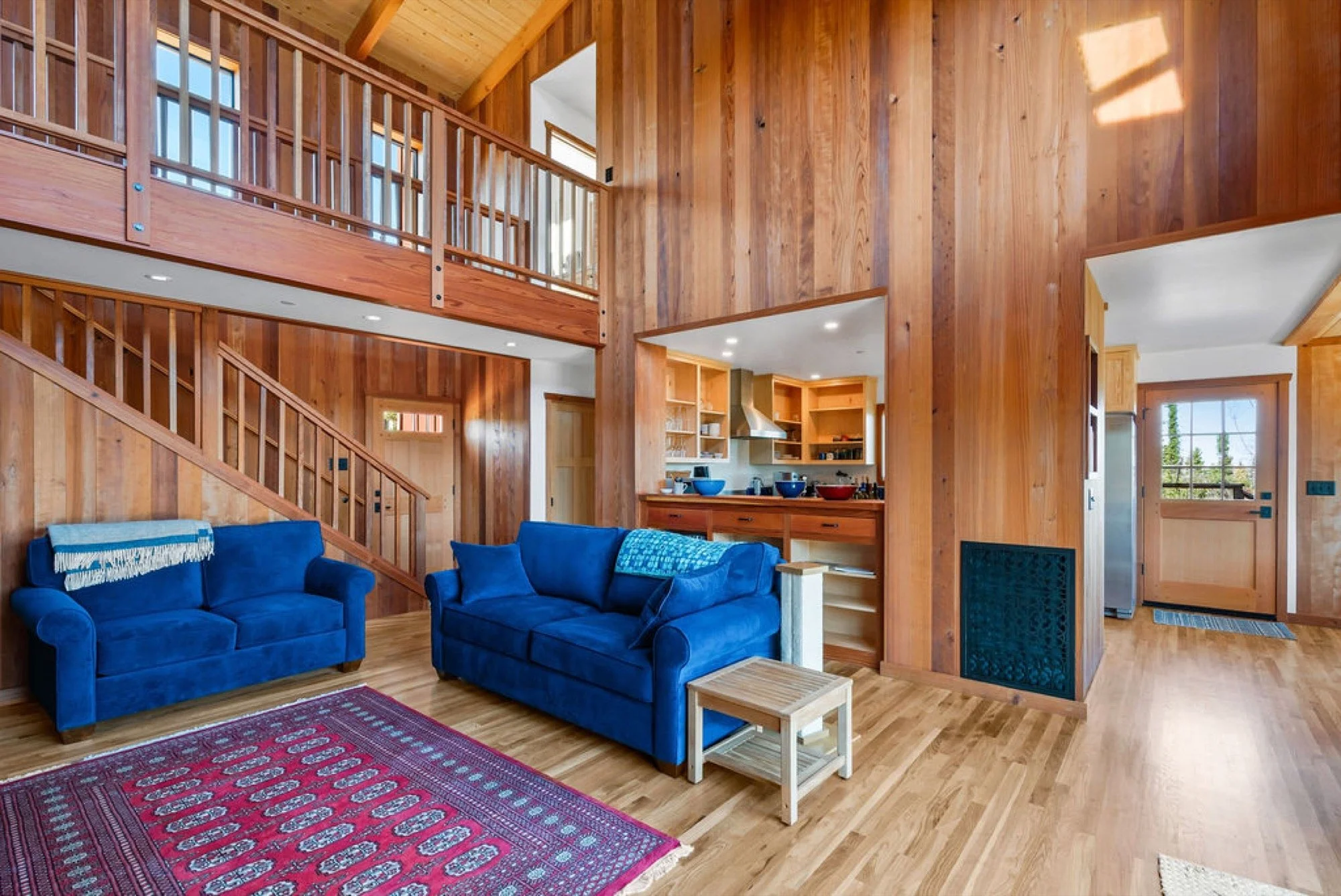 Interior of a wooden cabin living room with two blue sofas, a red patterned rug, an open kitchen with wooden shelves and blue bowls, a staircase leading to an upper level, and a door leading outside.