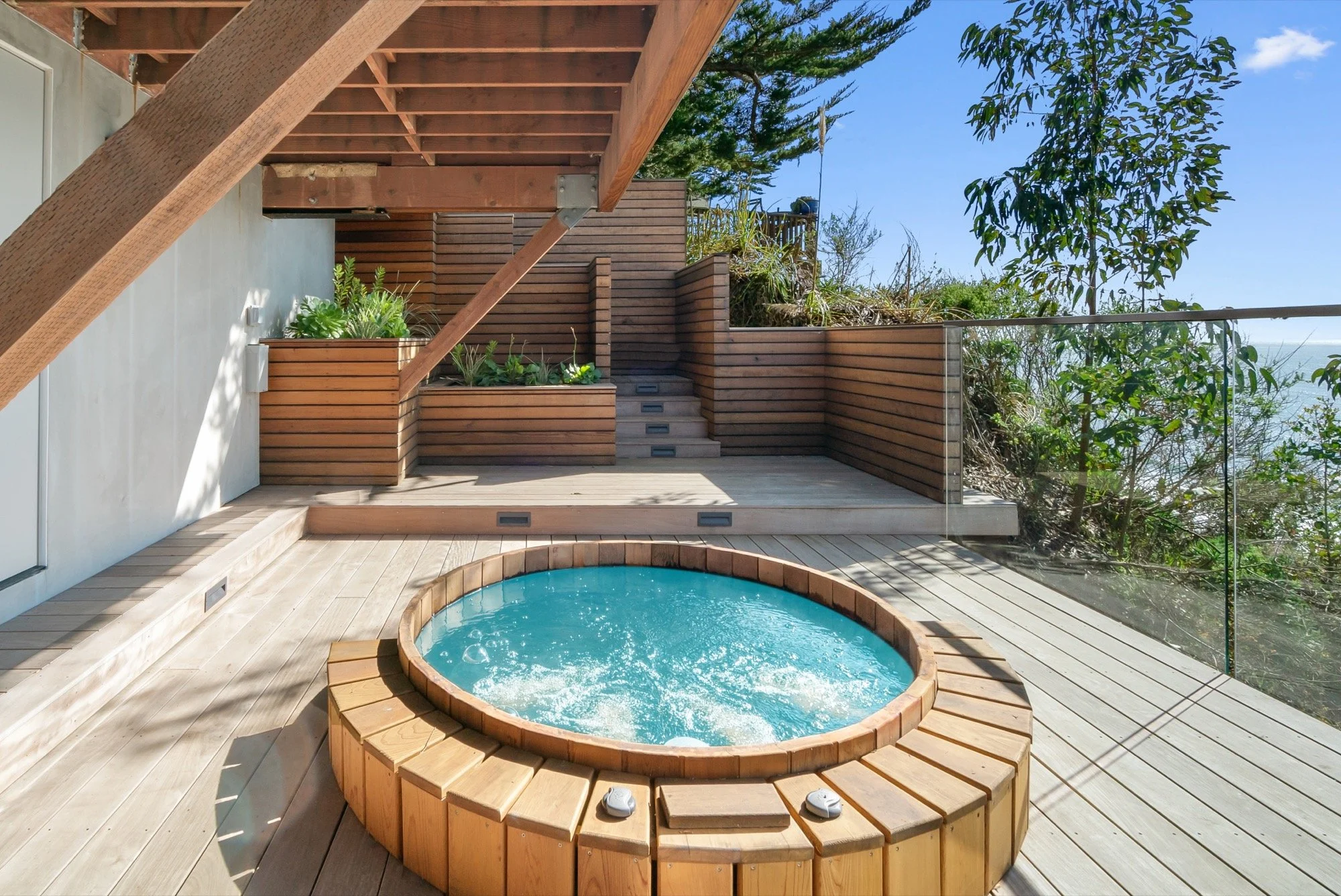 Outdoor wooden deck area with a small round hot tub filled with bubbling water, surrounded by wooden planks, and green plants in a wooden planter box. There are stairs leading up to a higher level with additional plants and a glass railing on the sid