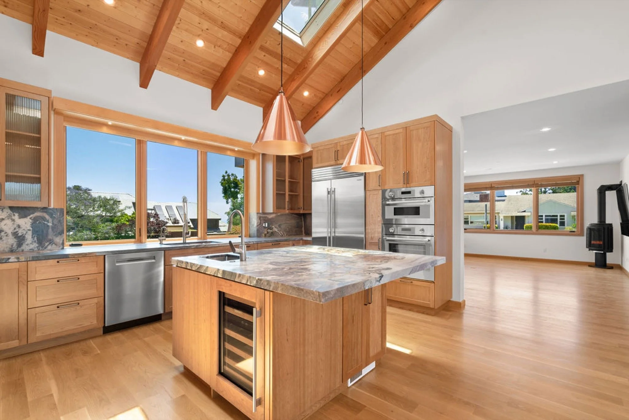 Modern kitchen with wooden cabinets, marble countertops, and large windows with a view of trees and neighboring houses.